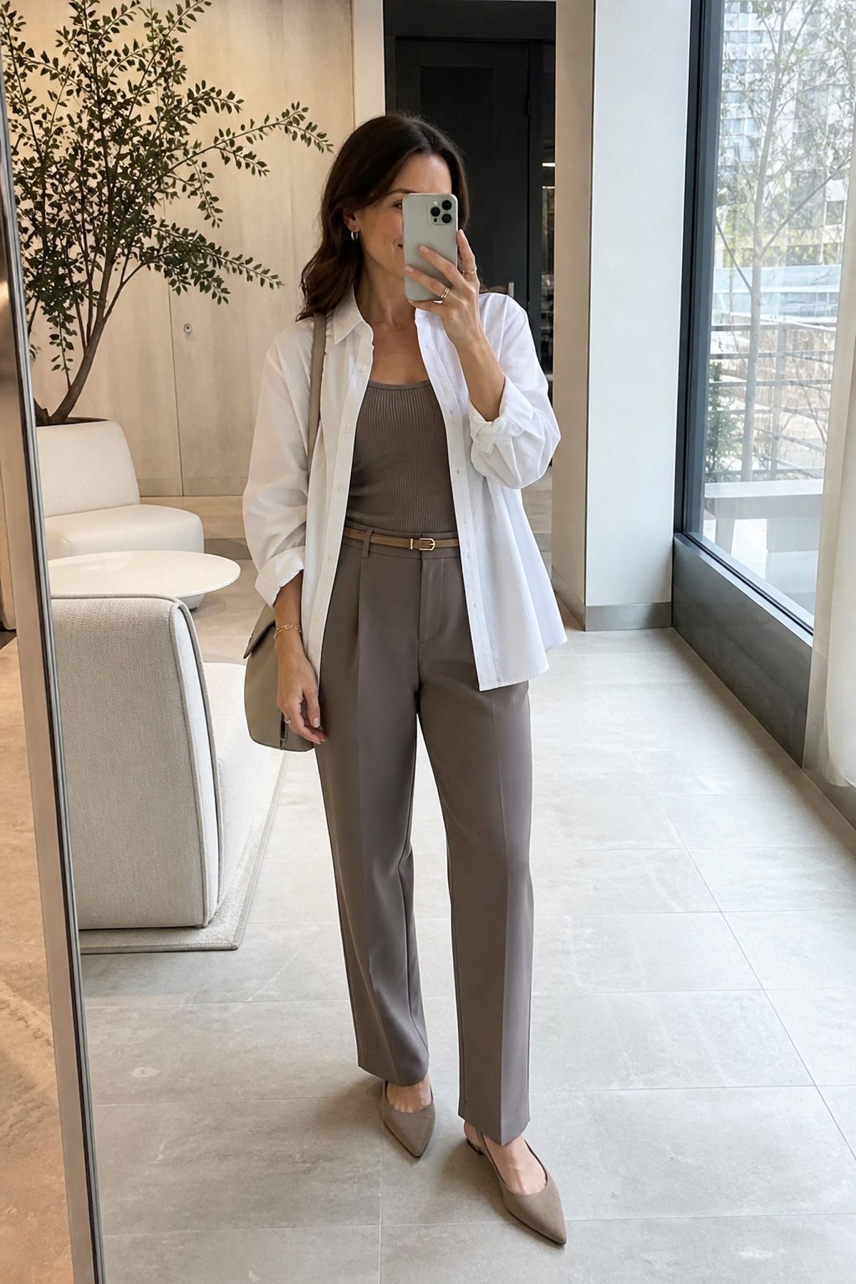 Mirror selfie of a woman in an open white button-down shirt over a taupe tank and taupe belted trousers with nude pointed heels in a bright office lobby