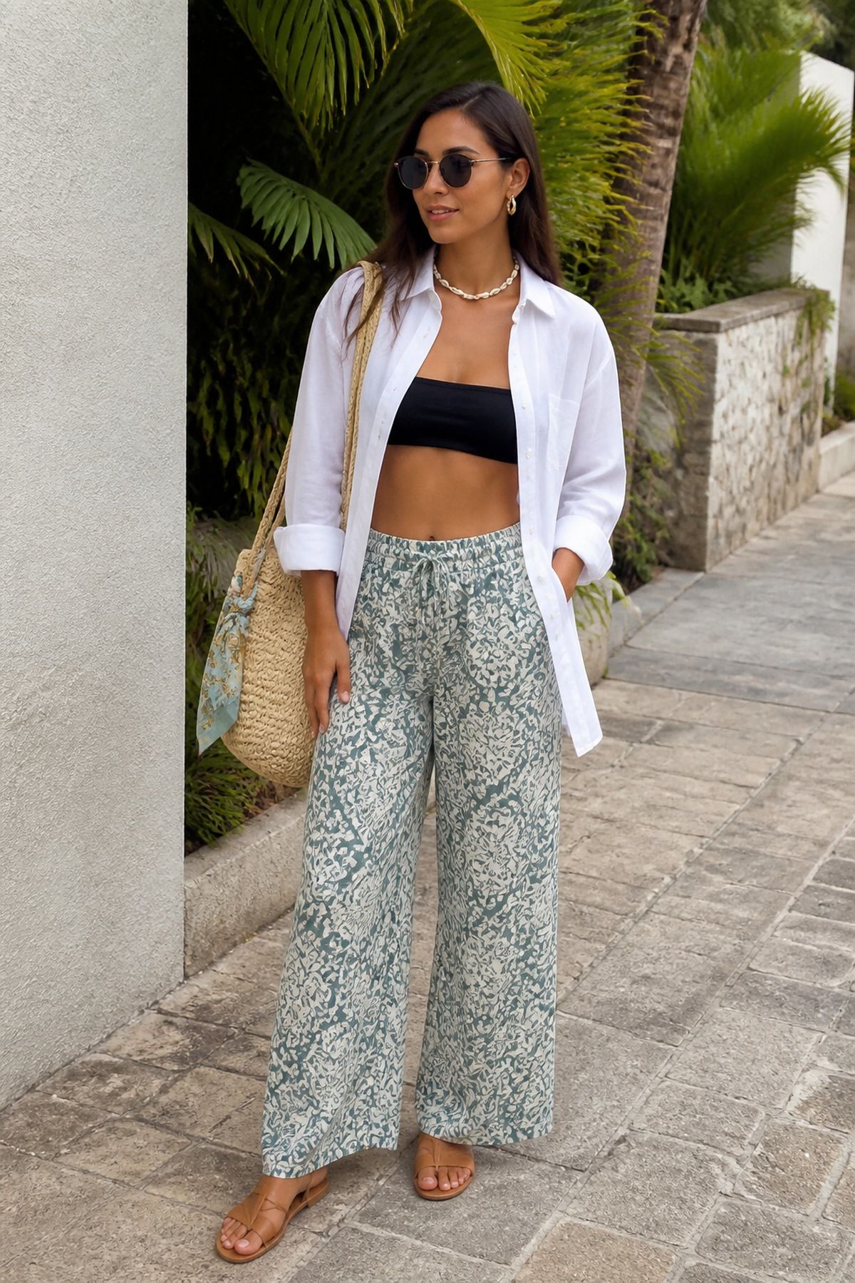 Woman in a black bandeau bikini top, an open white shirt and green and white paisley wide pants with flat sandals and a straw bag under palm trees