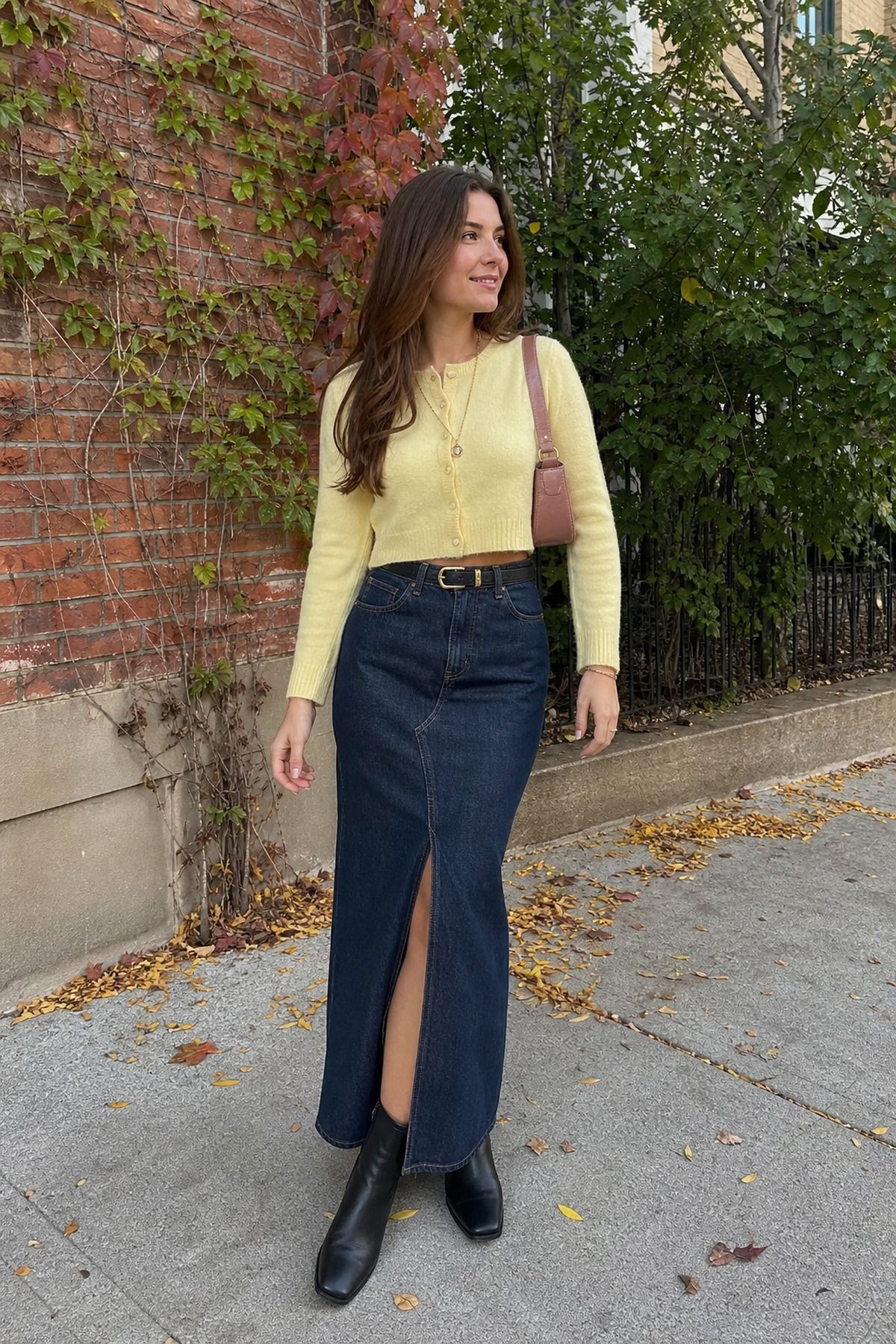 Woman in a butter yellow cropped cardigan with a dark denim maxi skirt with a front slit and black ankle boots on a leaf-covered sidewalk