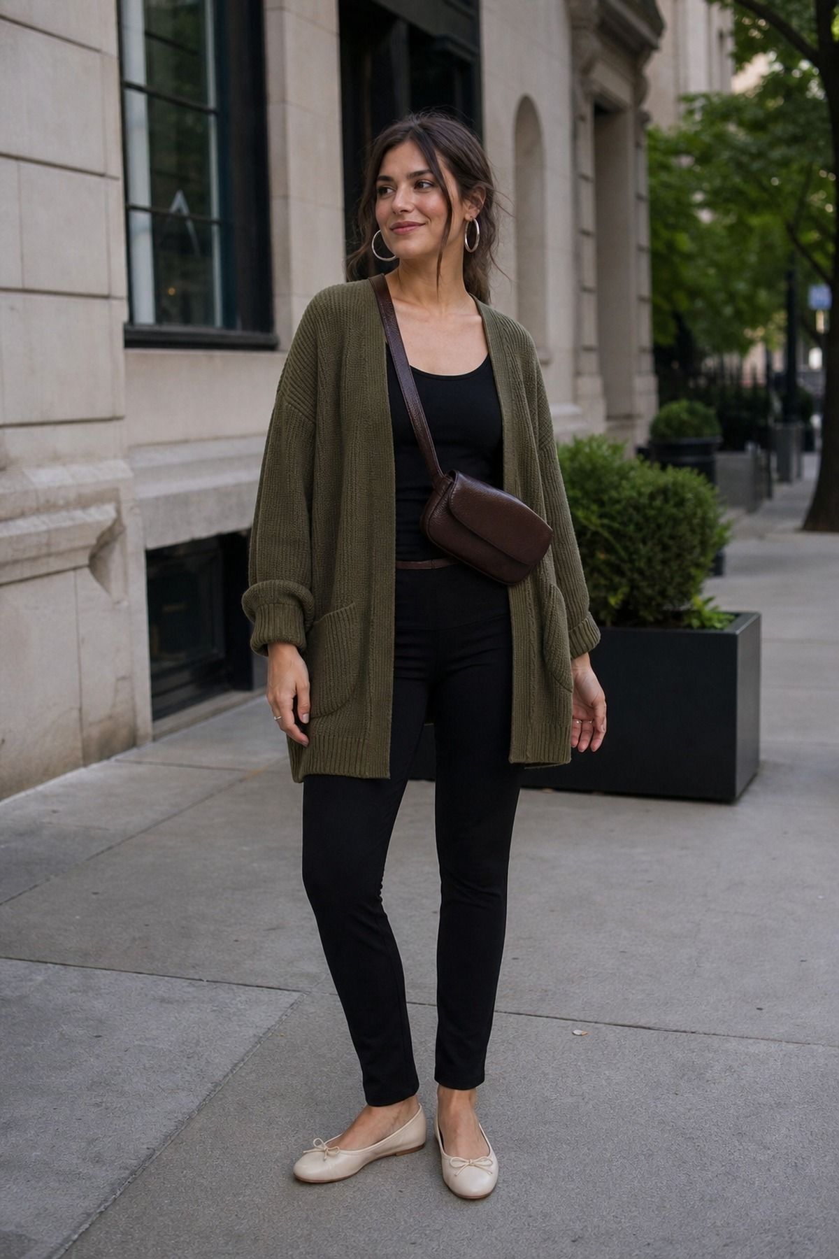 Woman in a long olive oversized cardigan over a black tank with black slim pants, cream ballet flats and a brown crossbody bag