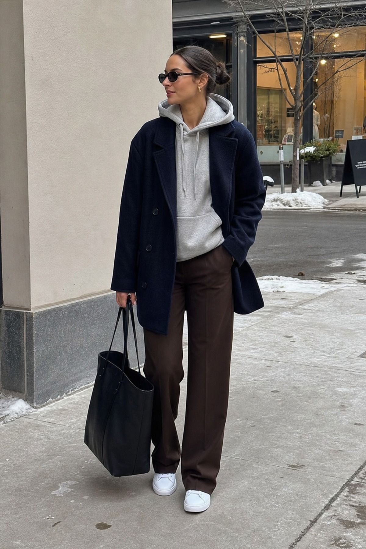 Woman in a navy wool blazer-style jacket over a gray hoodie with chocolate brown trousers, white sneakers and a black tote on a snowy street