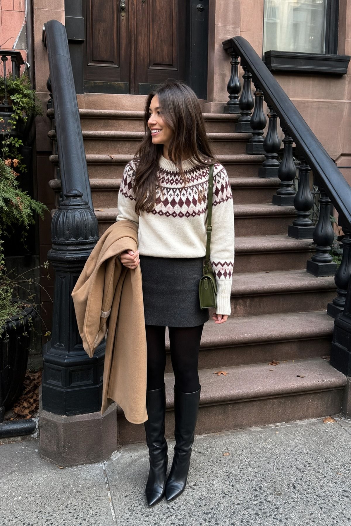 Woman in a cream and burgundy Fair Isle sweater with a charcoal wool mini skirt, black tights and black knee-high boots holding a camel coat by brownstone steps