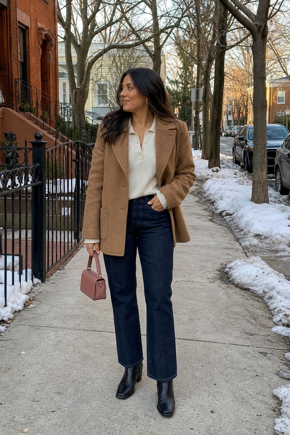 Woman in a cream knit polo under a camel jacket with dark straight jeans, black ankle boots and a pink bag on a snowy sidewalk