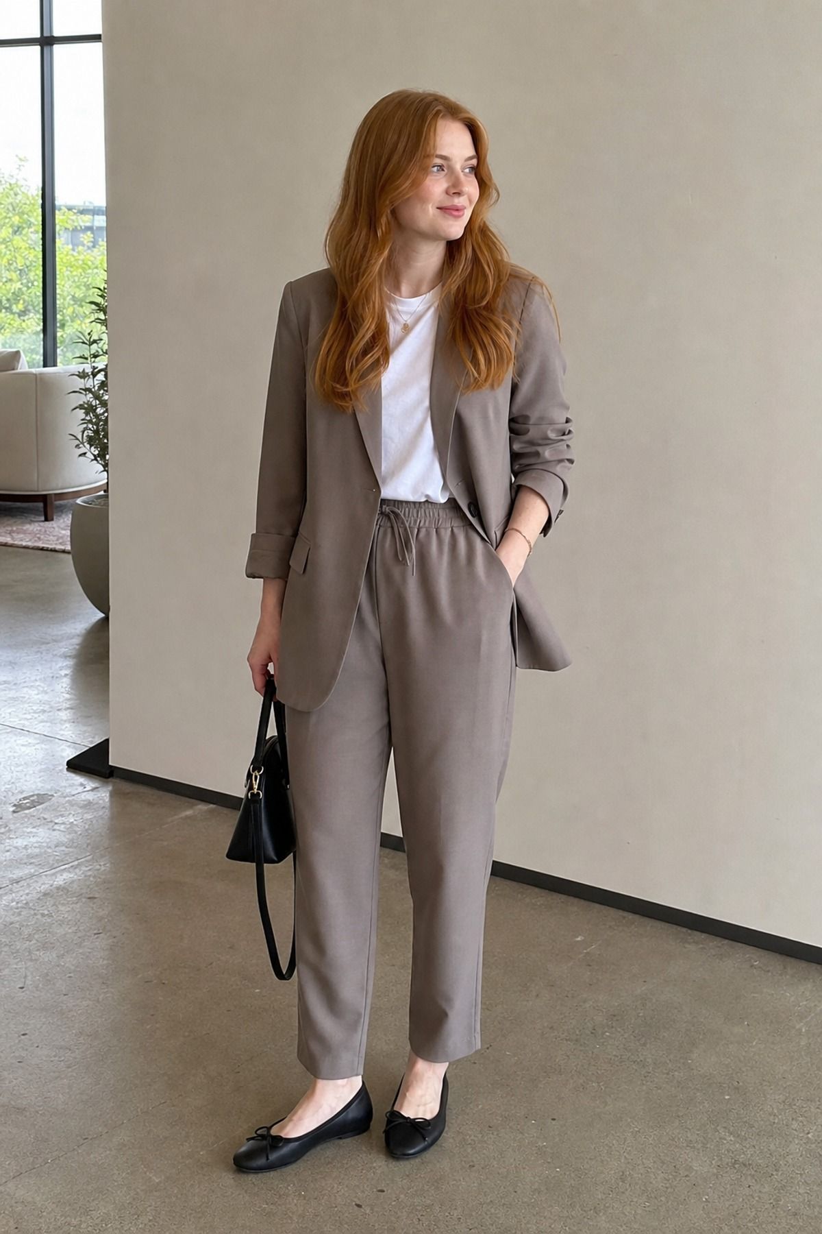 Woman in a taupe-gray relaxed suit with a white tee, black ballet flats and a black handbag against a pale wall