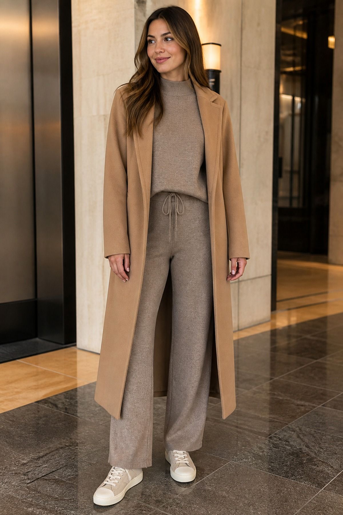 Woman in a taupe knit set of sweater and drawstring pants under a long camel coat with cream sneakers in a lobby