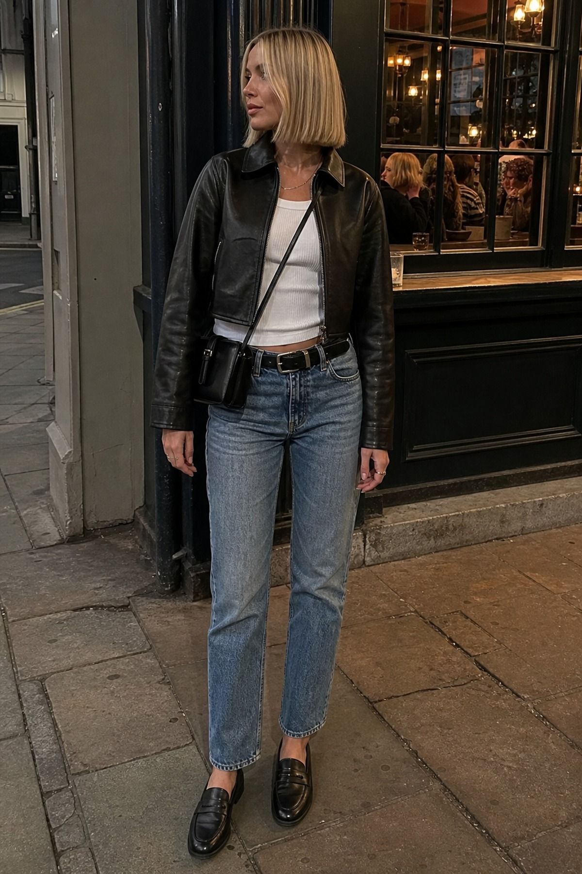 Woman in a black cropped leather jacket over a white tank with a black belt, light-wash straight jeans, black loafers and a black crossbody bag outside a pub