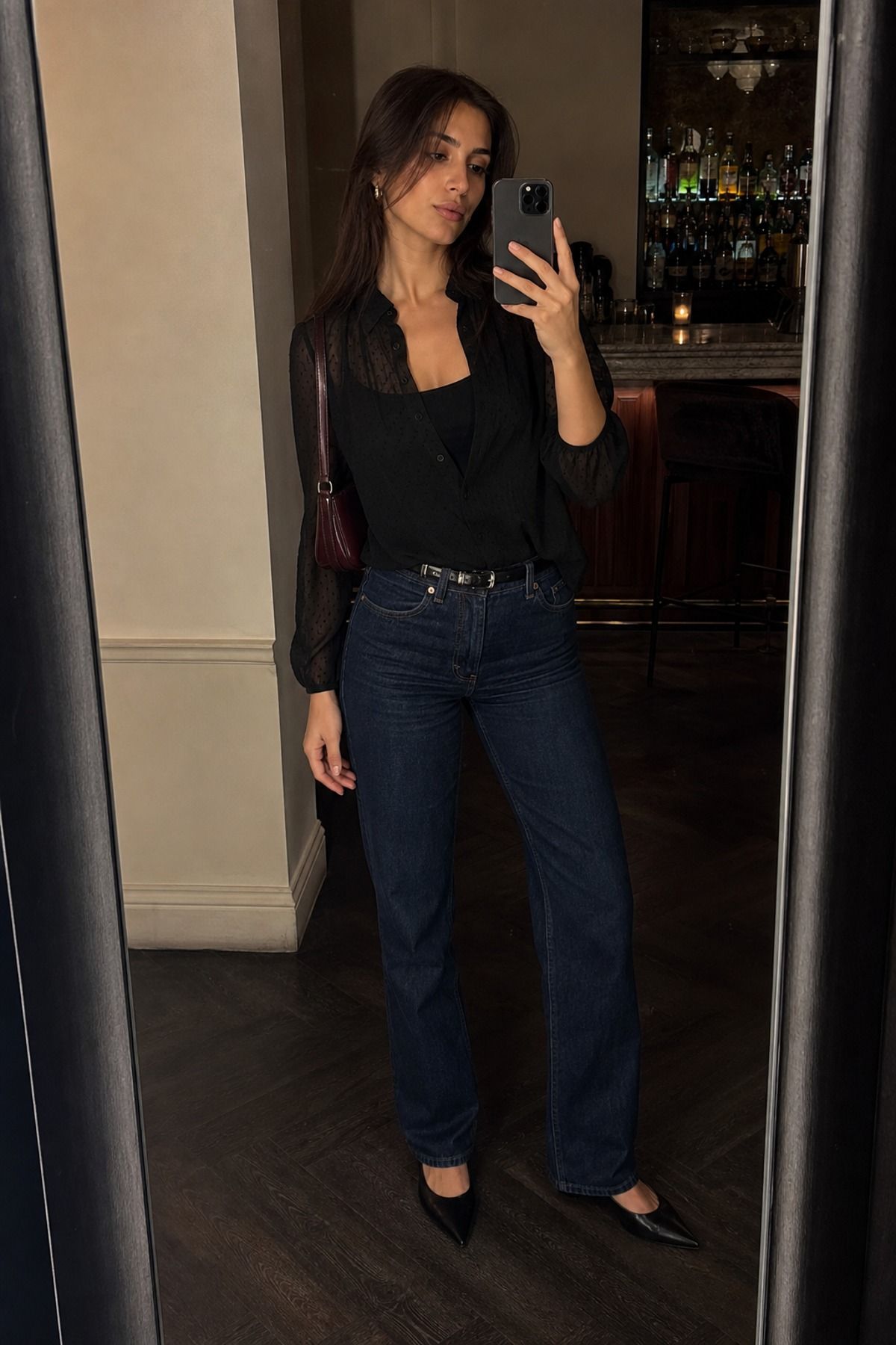 Mirror selfie of a woman in a black sheer blouse over a black cami with dark straight jeans, a black belt, black pointed shoes and a burgundy shoulder bag in a bar
