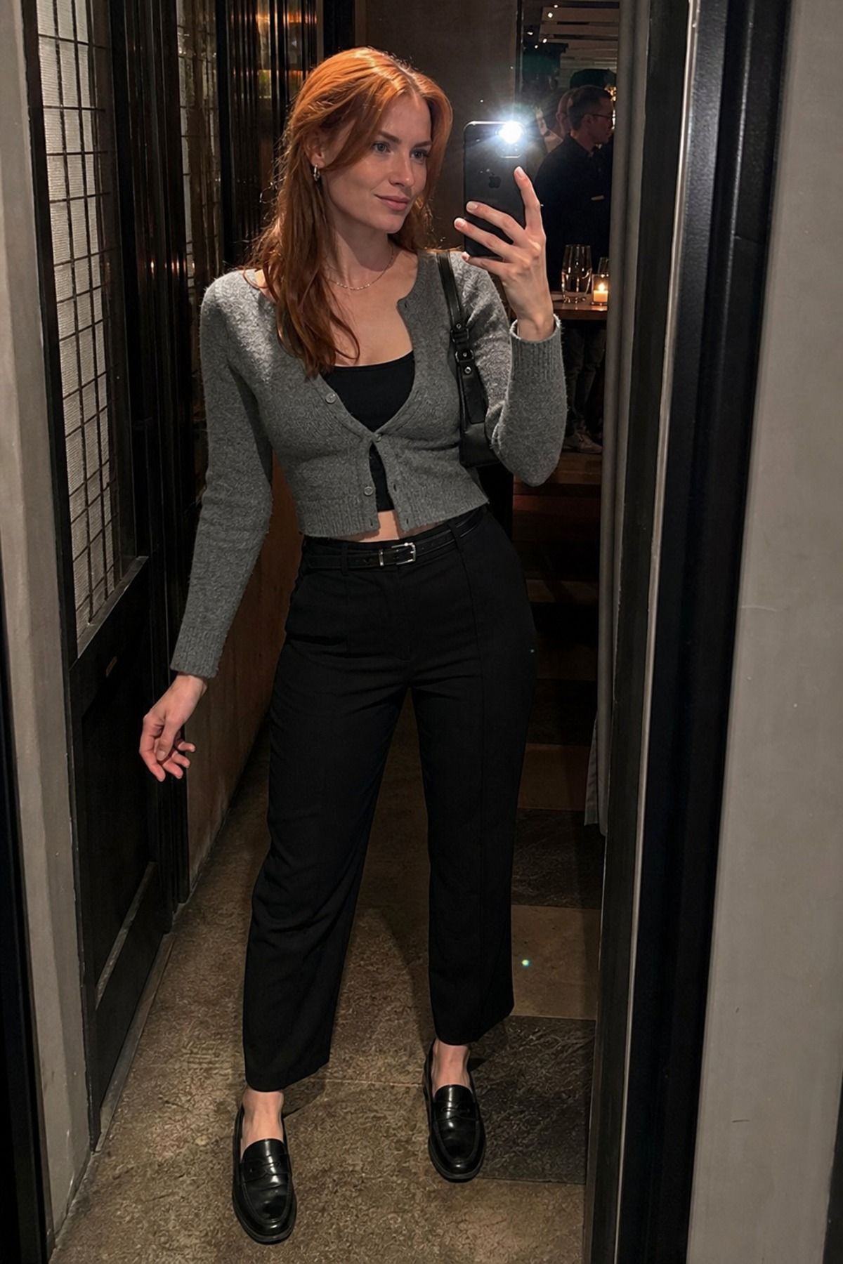 Mirror selfie of a woman in a gray cropped cardigan over a black top with black trousers, a black belt and black loafers in a bar entrance
