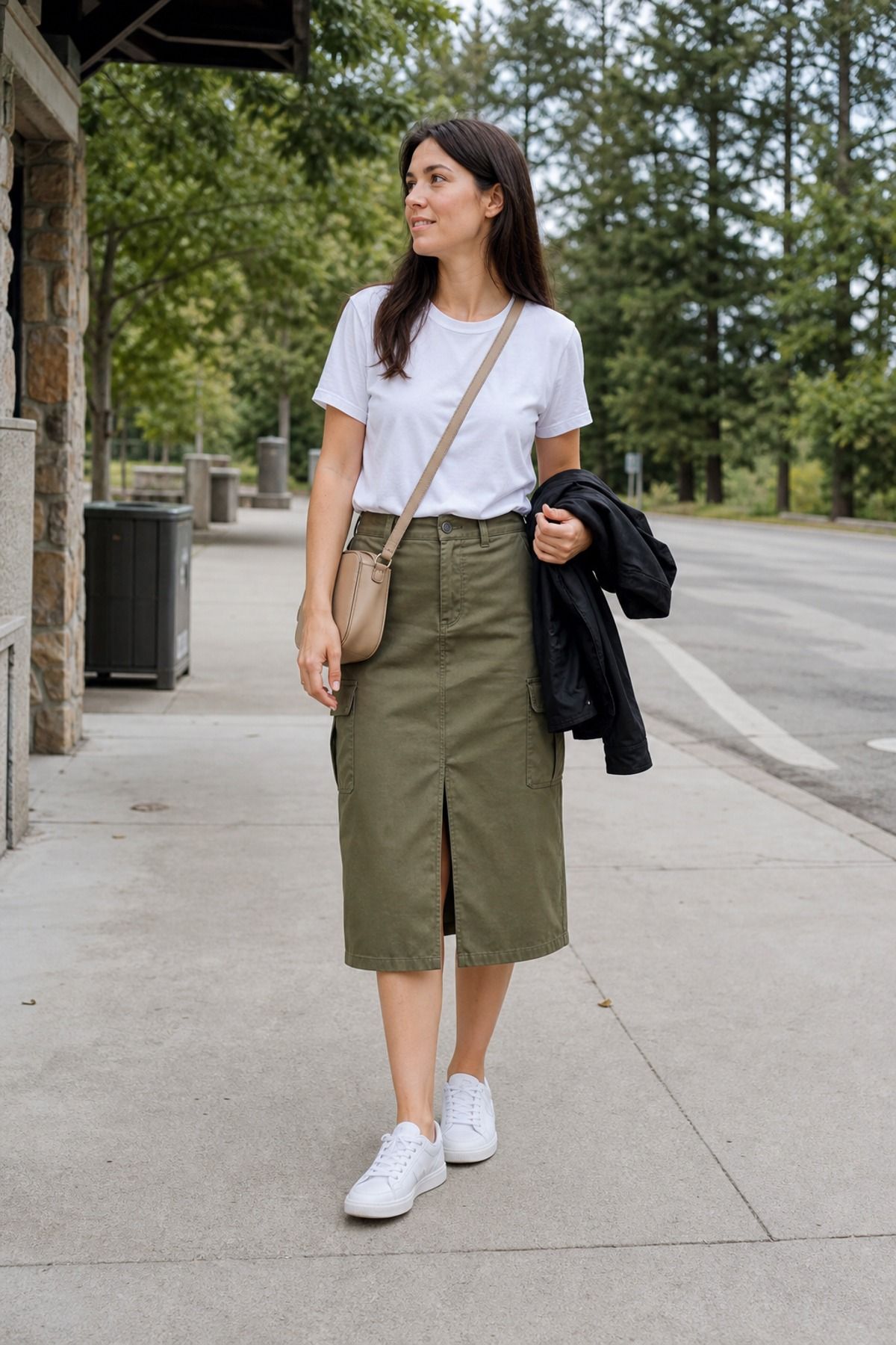 Woman in an olive midi cargo skirt with a front slit, a white cotton tee, white sneakers and a tan crossbody bag with a black jacket over her arm on a tree-lined street