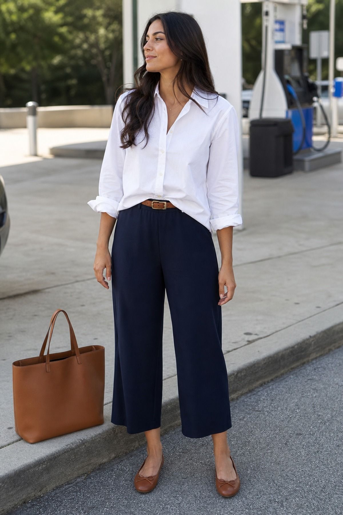 Woman in a white button-down shirt with rolled sleeves tucked into navy pull-on wide pants with a brown belt, brown flats and a cognac tote at a gas station