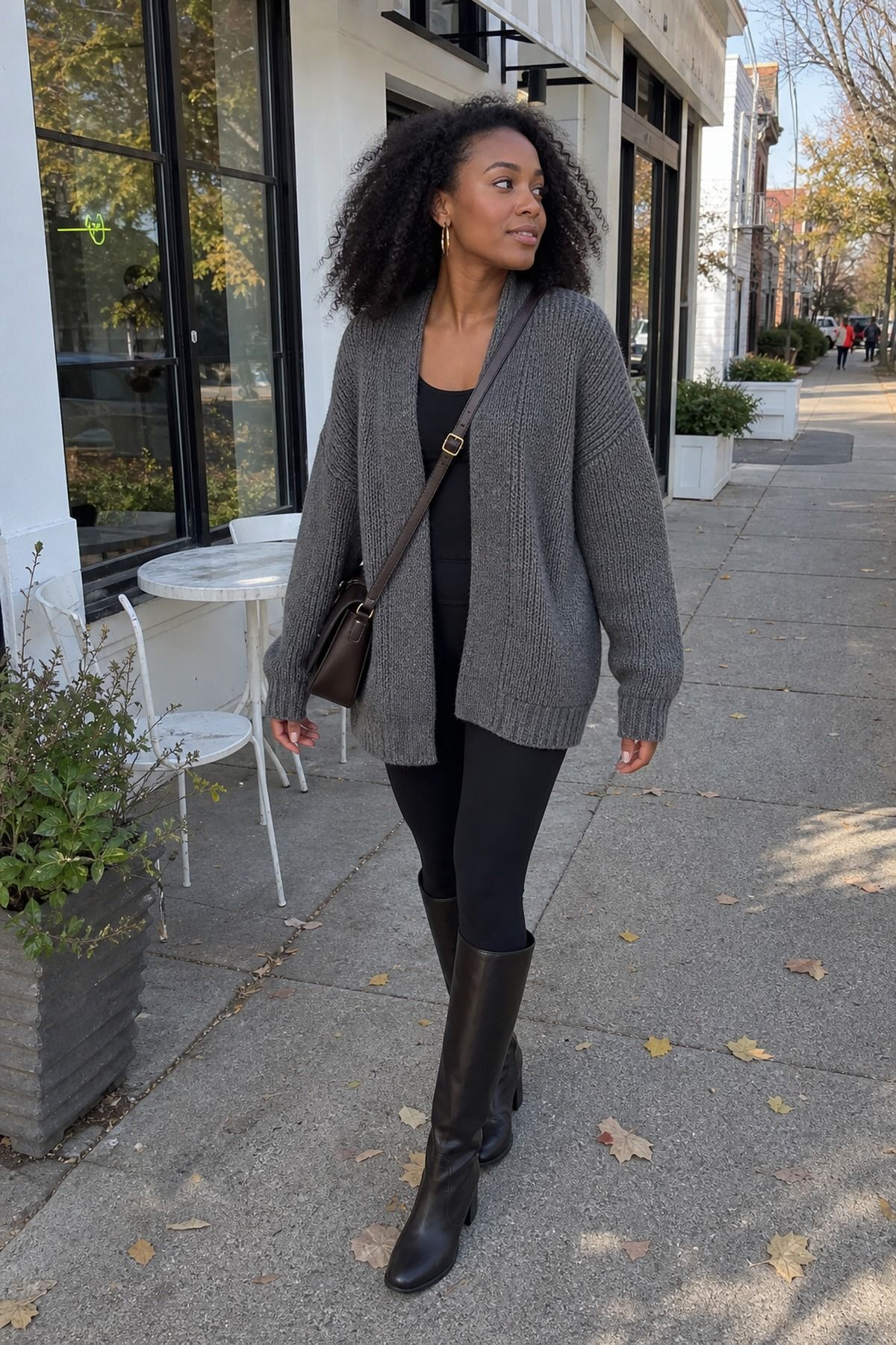 Woman in a gray chunky oversized cardigan over a black top with black leggings, black knee-high boots and a brown crossbody bag on a fall street