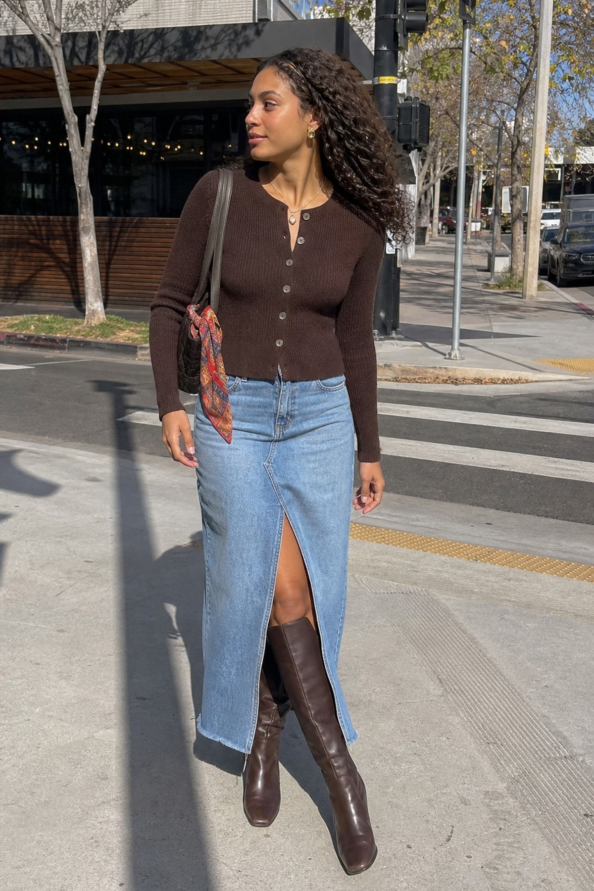 Woman in a chocolate brown fitted ribbed cardigan with a denim midi skirt with a front slit and brown knee-high boots crossing a street