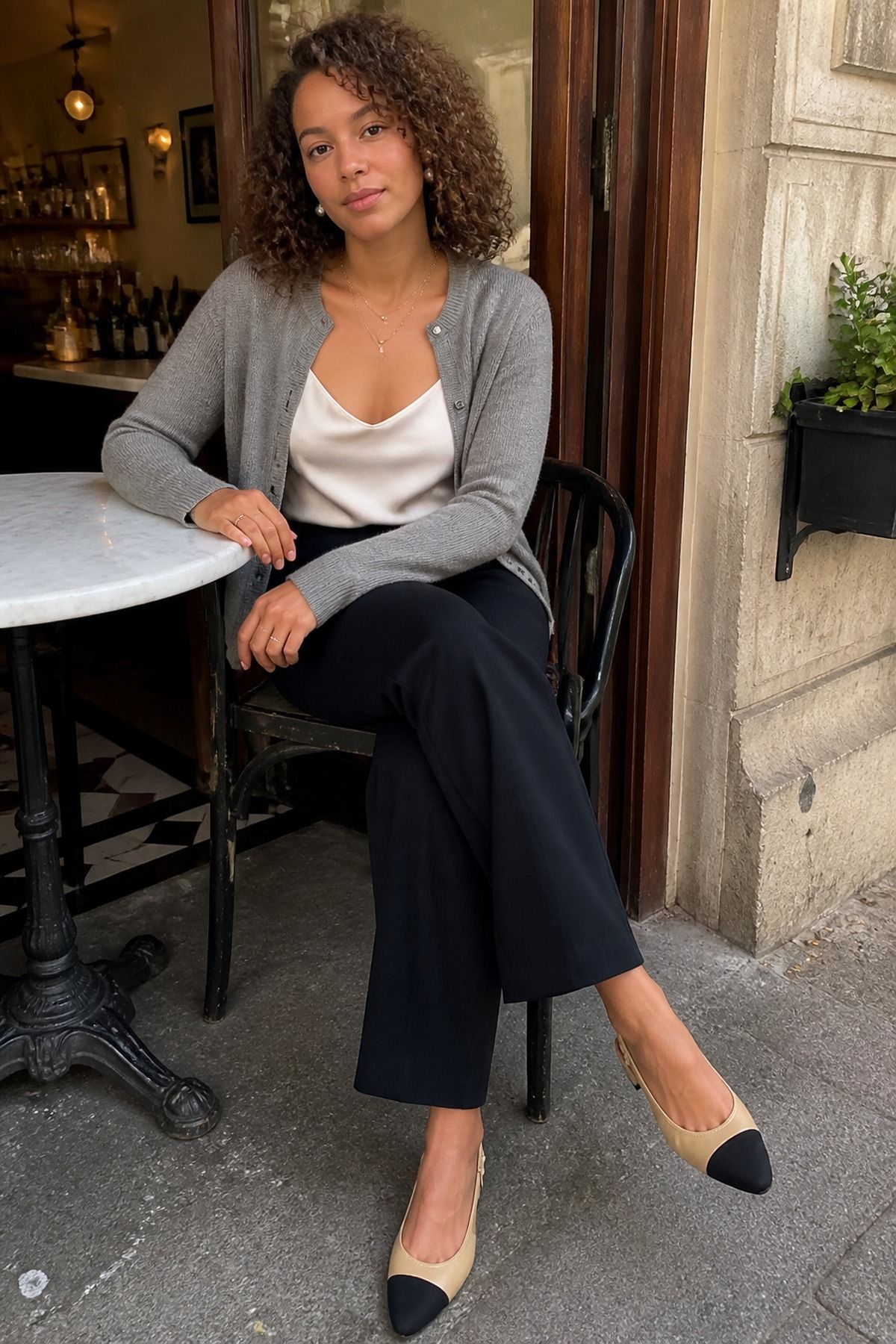 Woman in a gray fine-knit cardigan worn open over an ivory satin cami with black wide trousers and beige cap-toe slingbacks at a cafe table