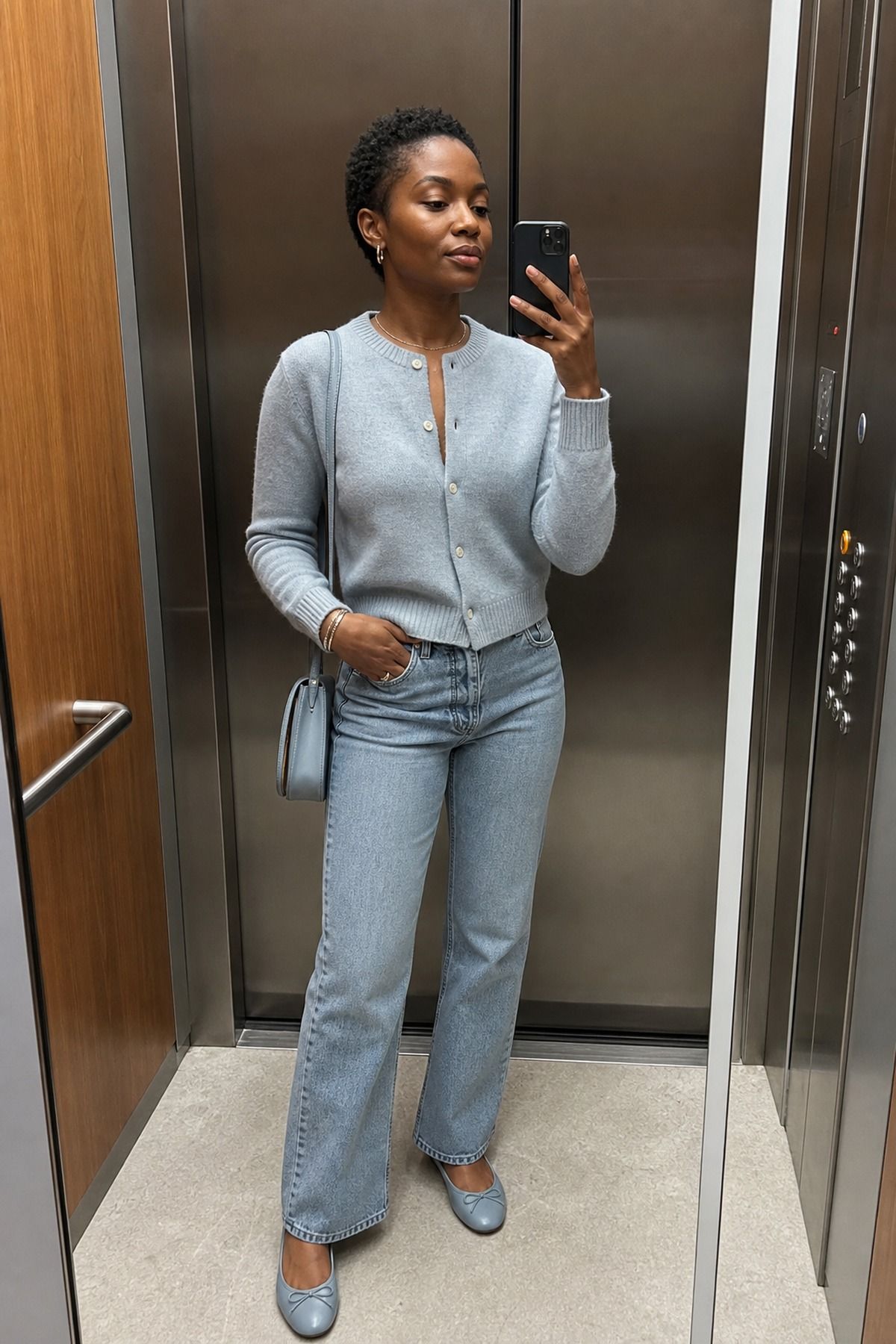 Elevator mirror selfie of a woman in a pale blue cropped cardigan with light-wash jeans, pale blue ballet flats and a blue shoulder bag