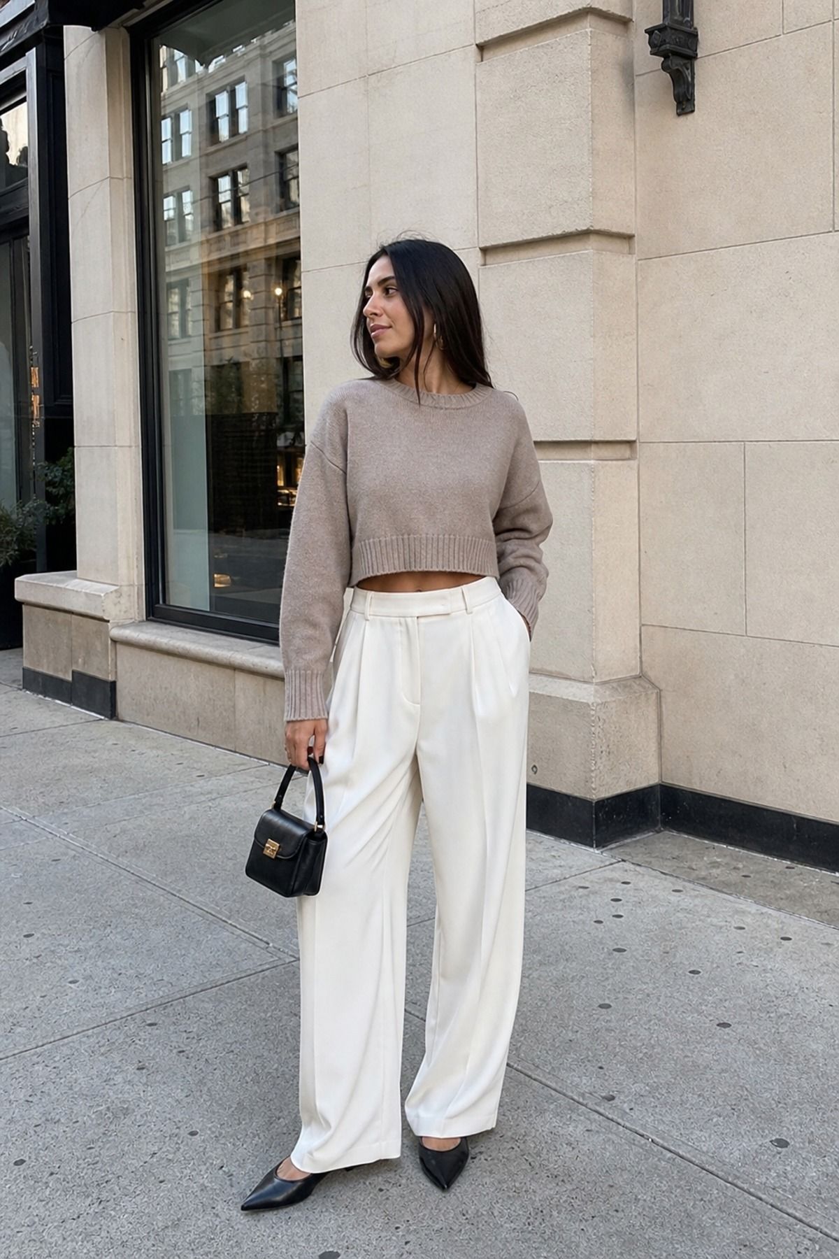 Woman in a taupe cropped sweater, cream pleated wide-leg trousers, black pointed slingback flats and a small black top-handle bag on a city street