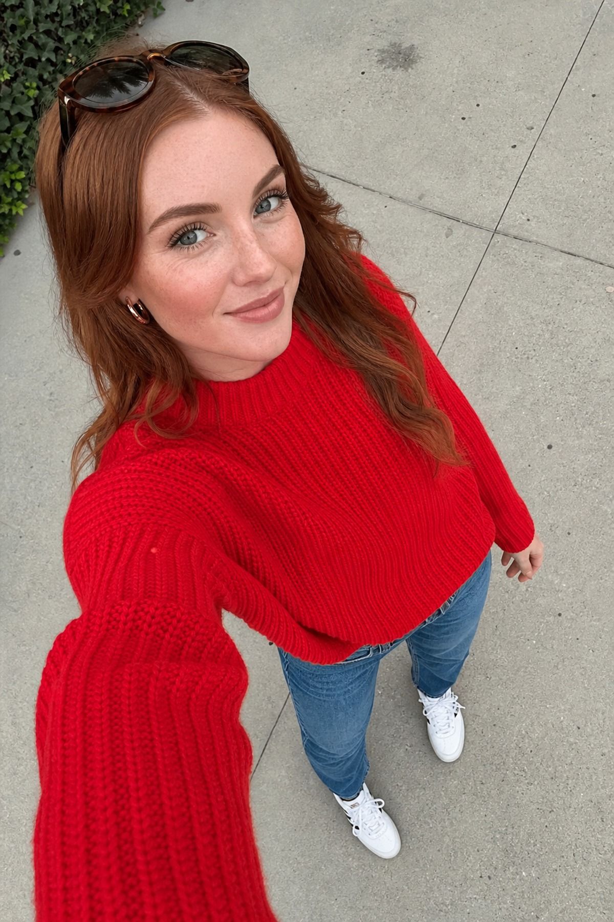 Selfie from above of a woman in a tomato red ribbed sweater, light blue jeans and white sneakers with sunglasses on her head