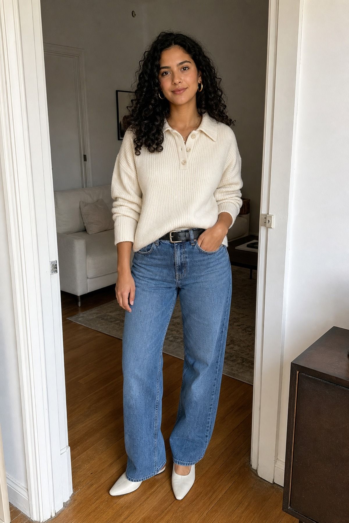 Woman in a cream knit polo sweater with relaxed mid-blue jeans and white pointed flats in a living room