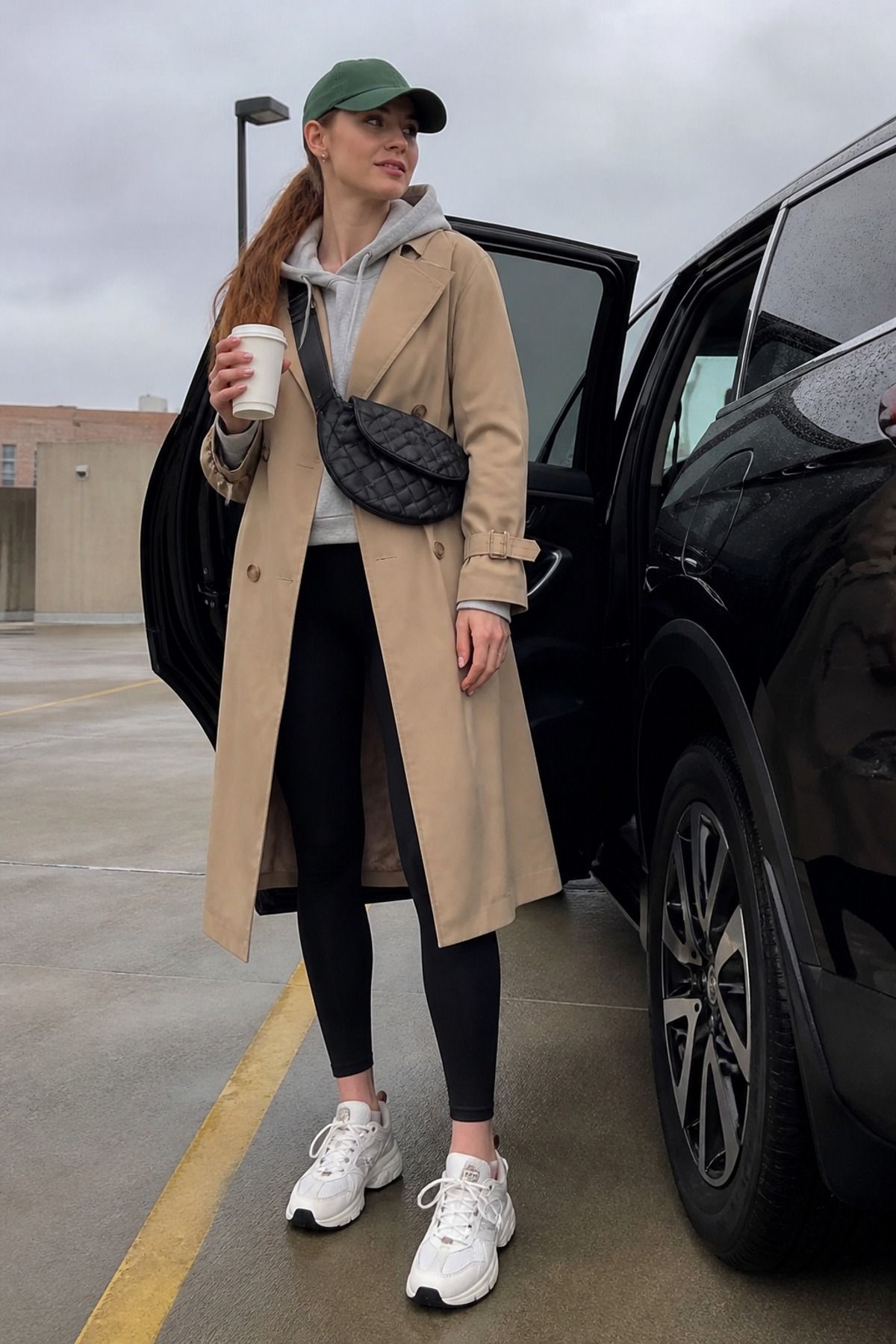 Woman in a tan trench coat over a gray hoodie and black leggings with white sneakers, a green cap and a quilted belt bag by a car