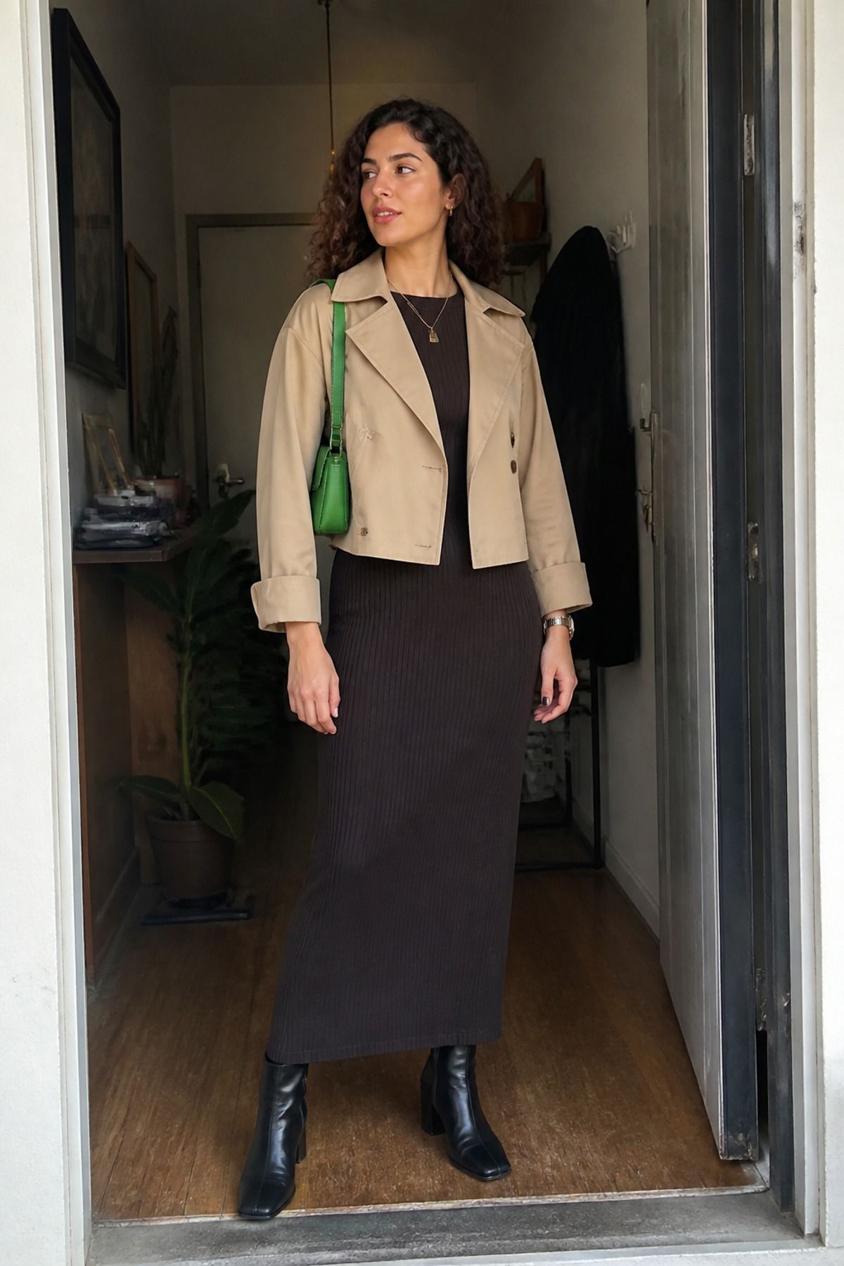 Woman in a cropped beige trench jacket over a dark ribbed column maxi dress with black ankle boots and a green shoulder bag