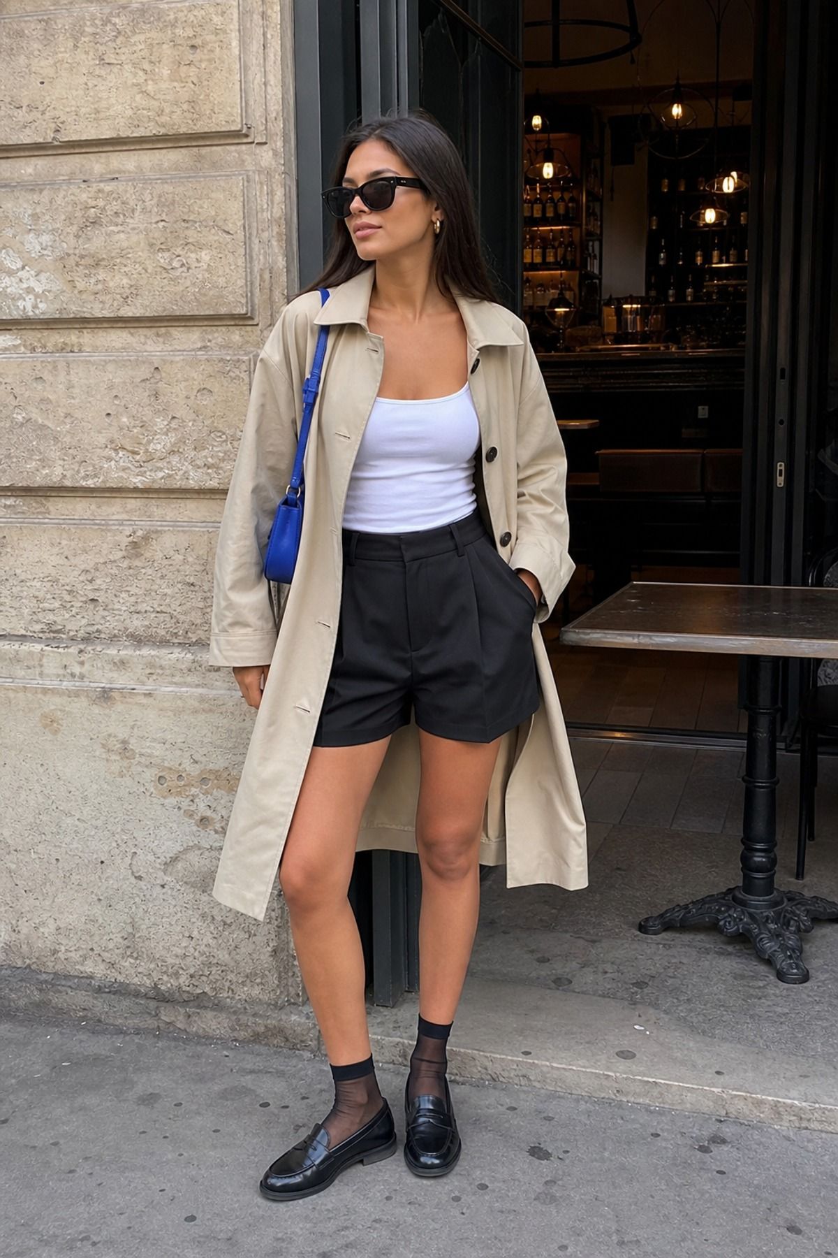 Woman in a beige trench coat, white tank top, black tailored shorts, sheer black socks and black loafers with a blue bag