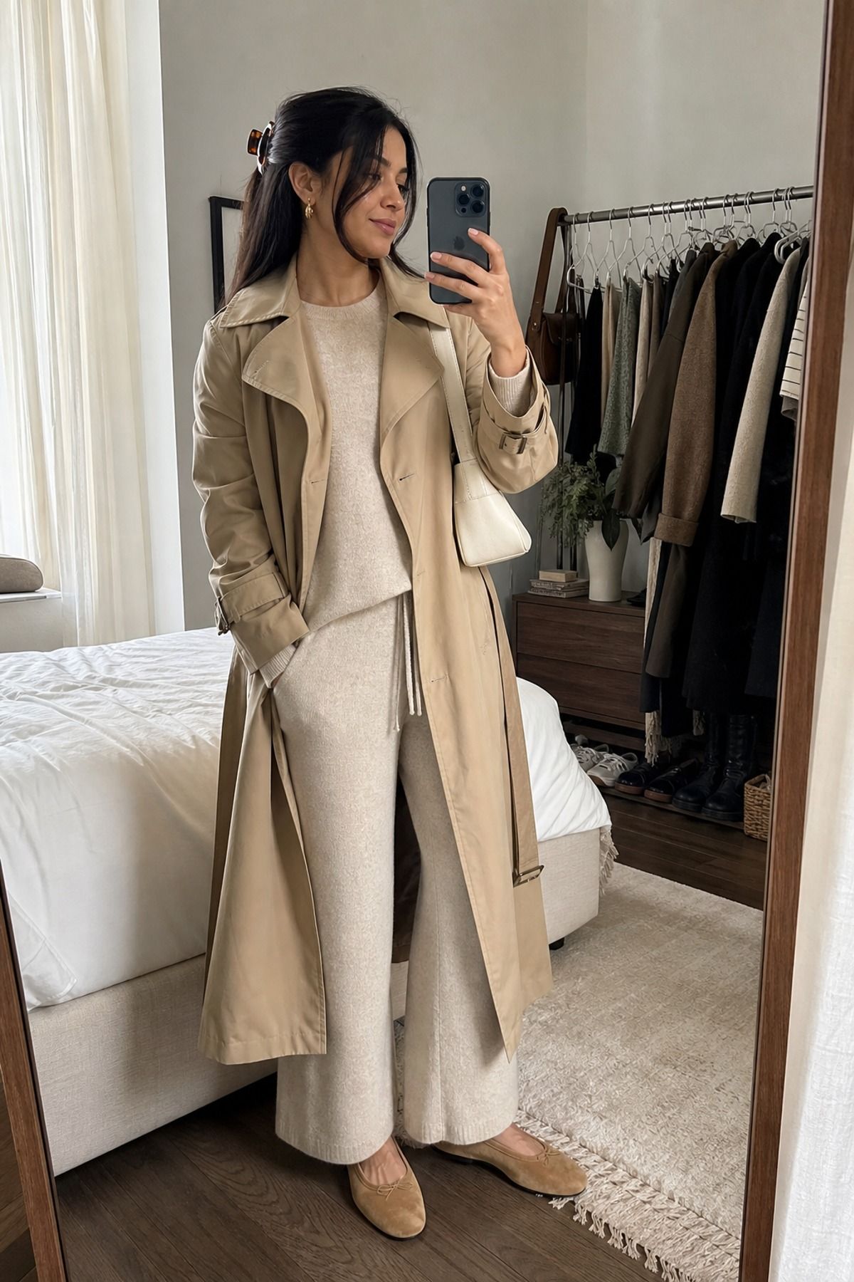 Woman in a beige trench coat over an oatmeal knit sweater and wide knit pants with tan ballet flats in a bedroom mirror selfie