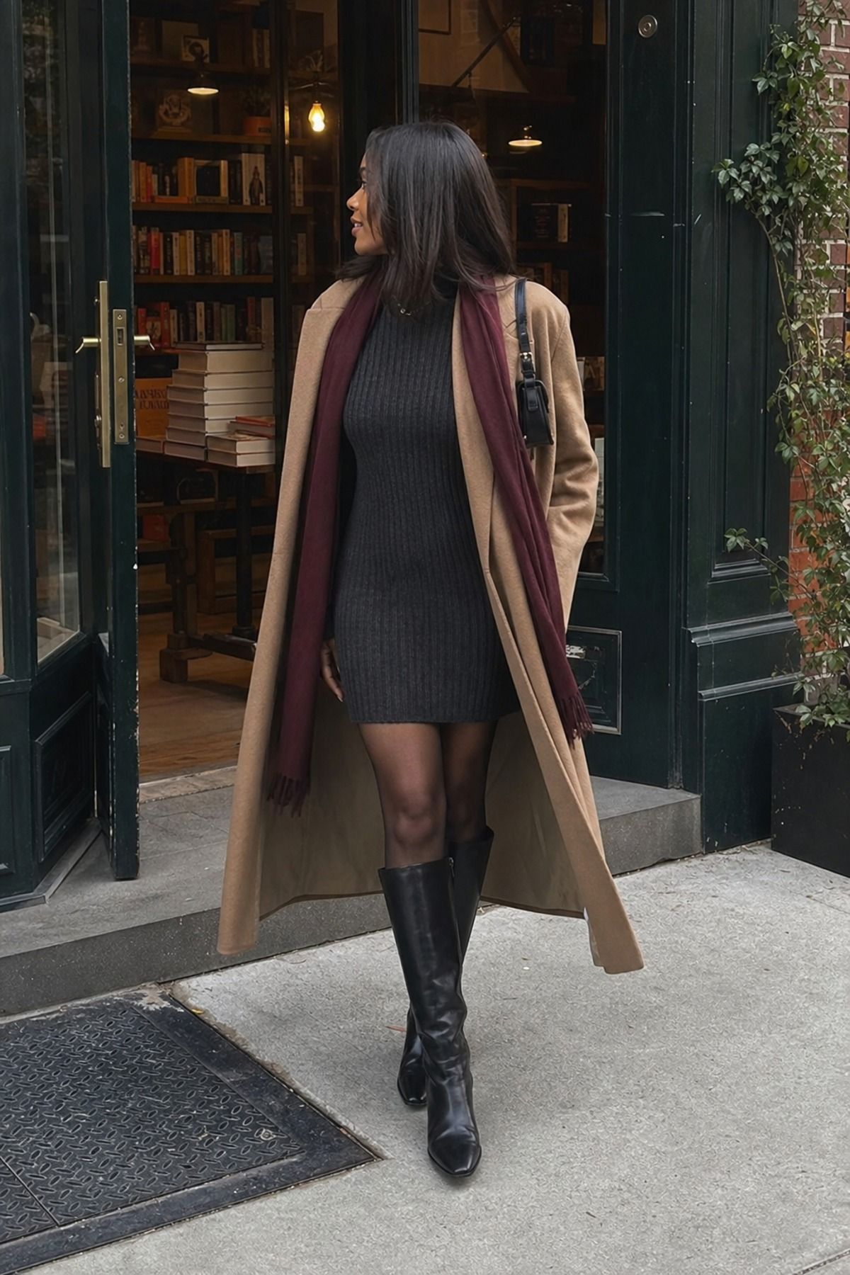 Woman in a charcoal ribbed sweater dress, long camel coat, burgundy scarf and tall black boots outside a bookshop