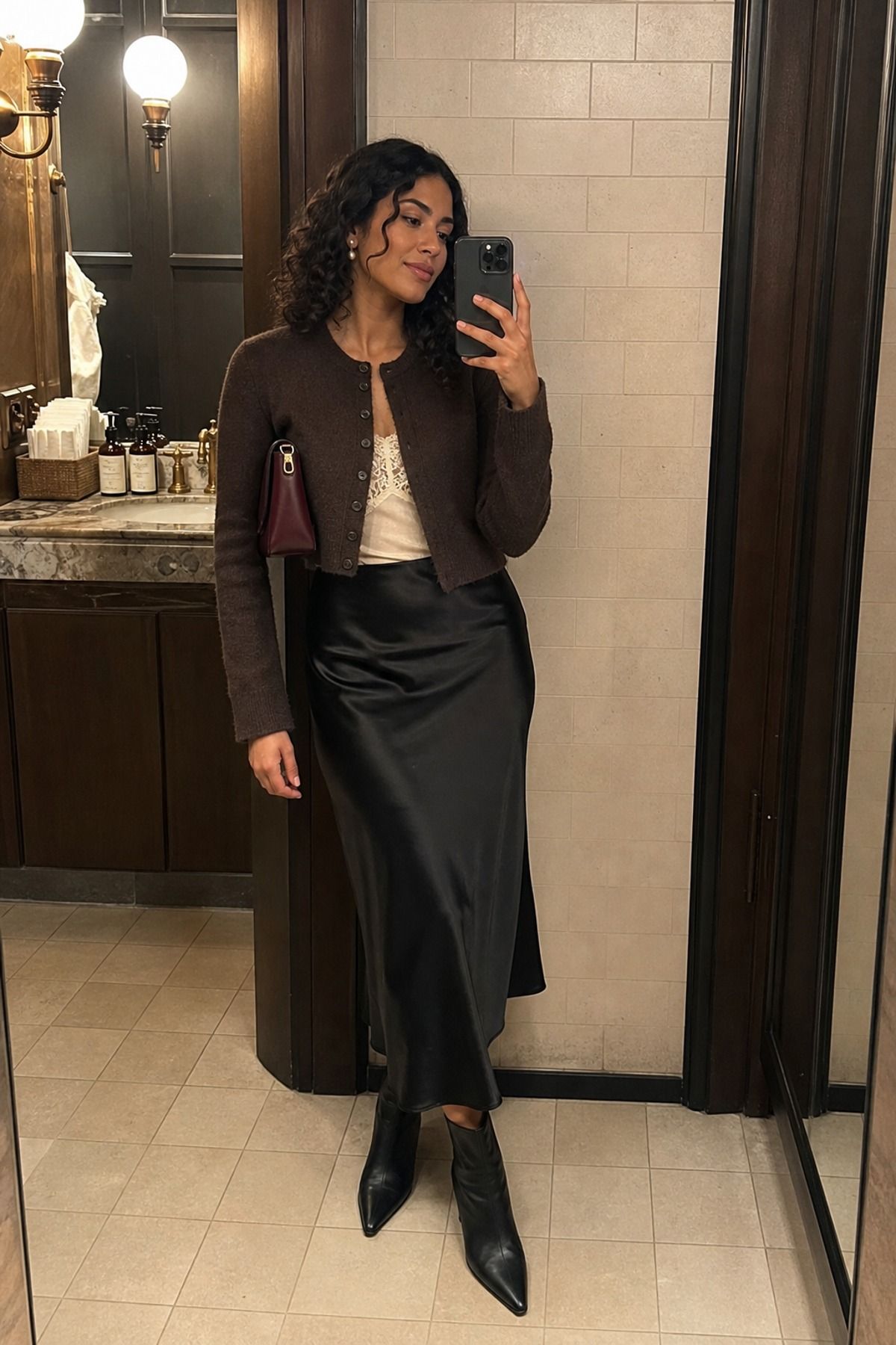 Woman in a black satin slip midi skirt and a brown cropped cardigan over a lace cami with black pointed ankle boots in a mirror selfie