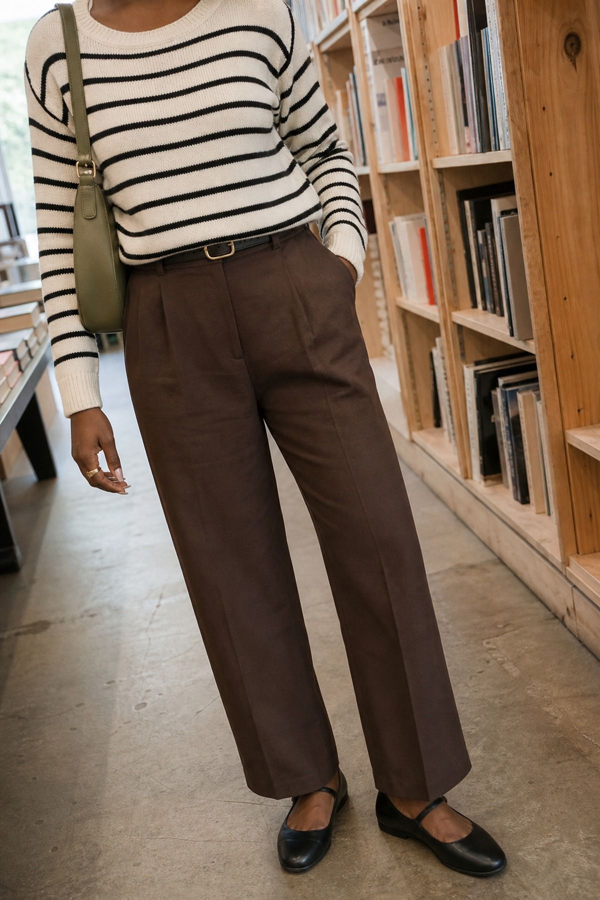 Woman in a cream and black striped knit and brown pleated trousers with black Mary Janes in a bookstore
