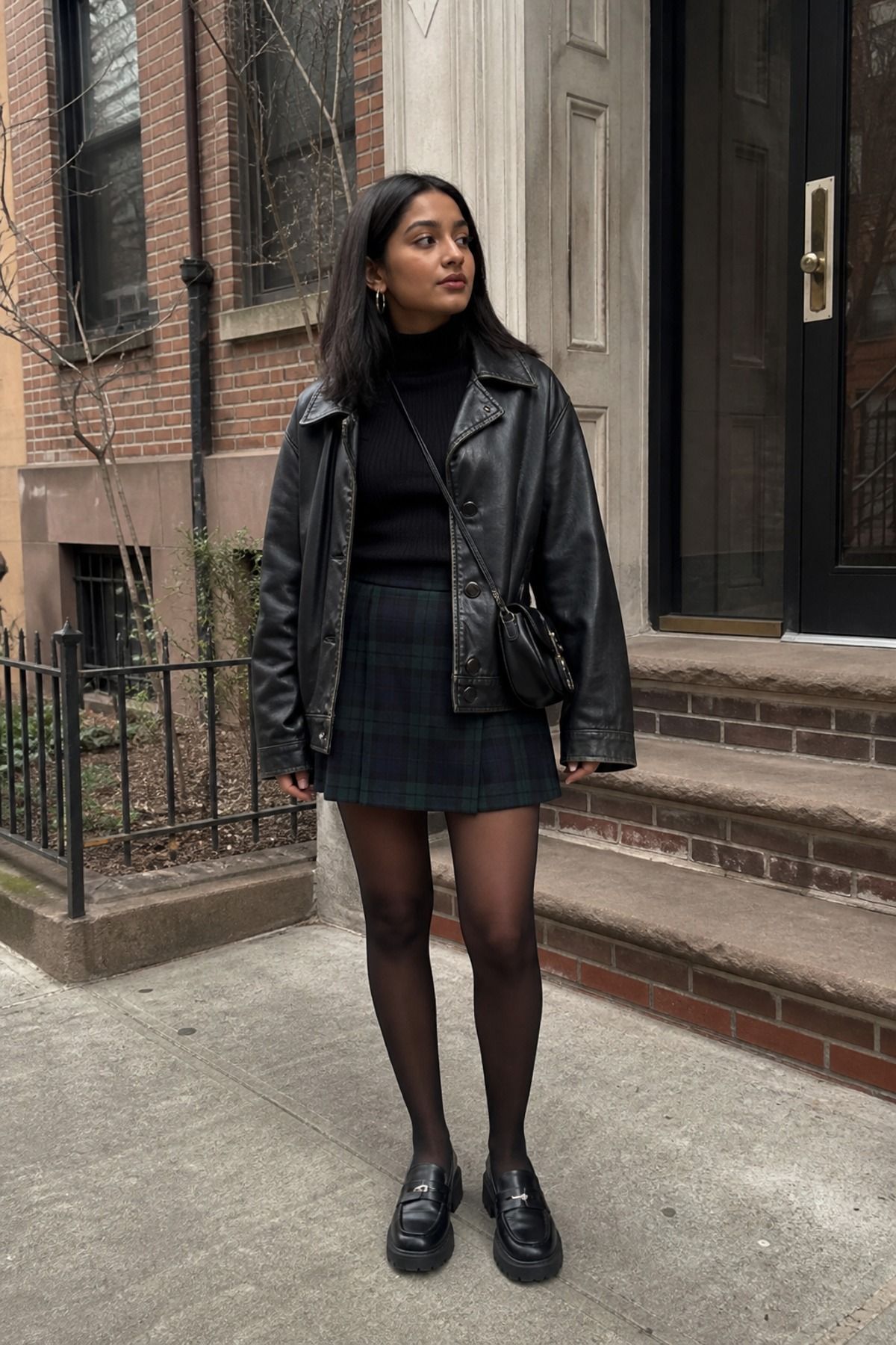 Woman in a black leather jacket, black turtleneck, dark plaid mini skirt, black tights and chunky loafers on a brownstone sidewalk