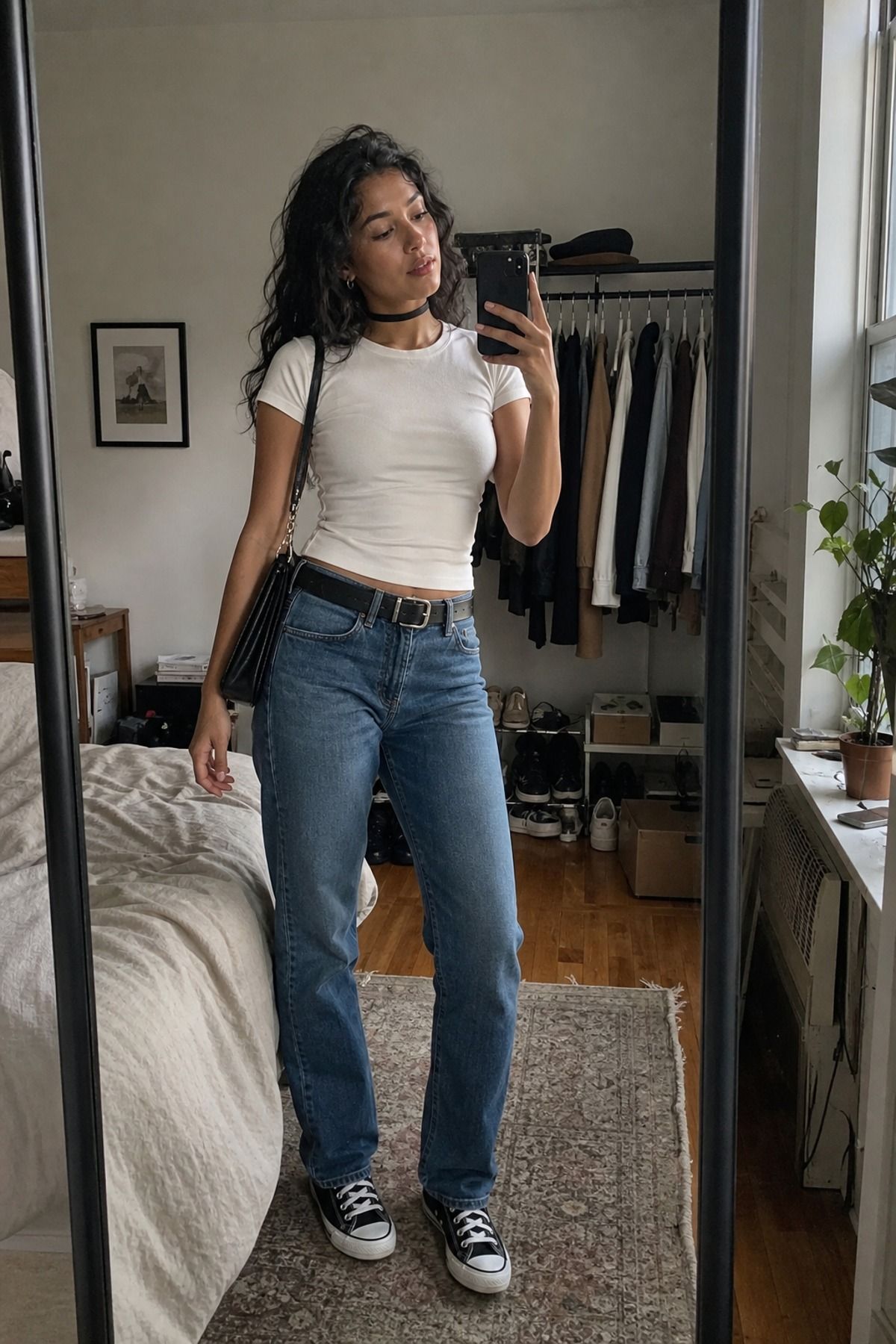 Mirror selfie of a woman in a black choker, fitted white baby tee, straight blue jeans with a black belt and black canvas high-top sneakers in a bedroom