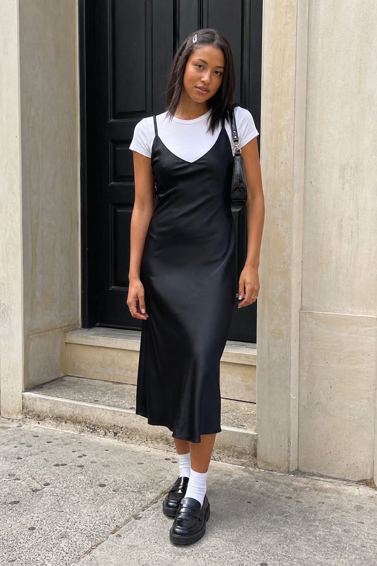 Woman in a black satin slip dress over a white baby tee with white socks and chunky black loafers in front of a black door