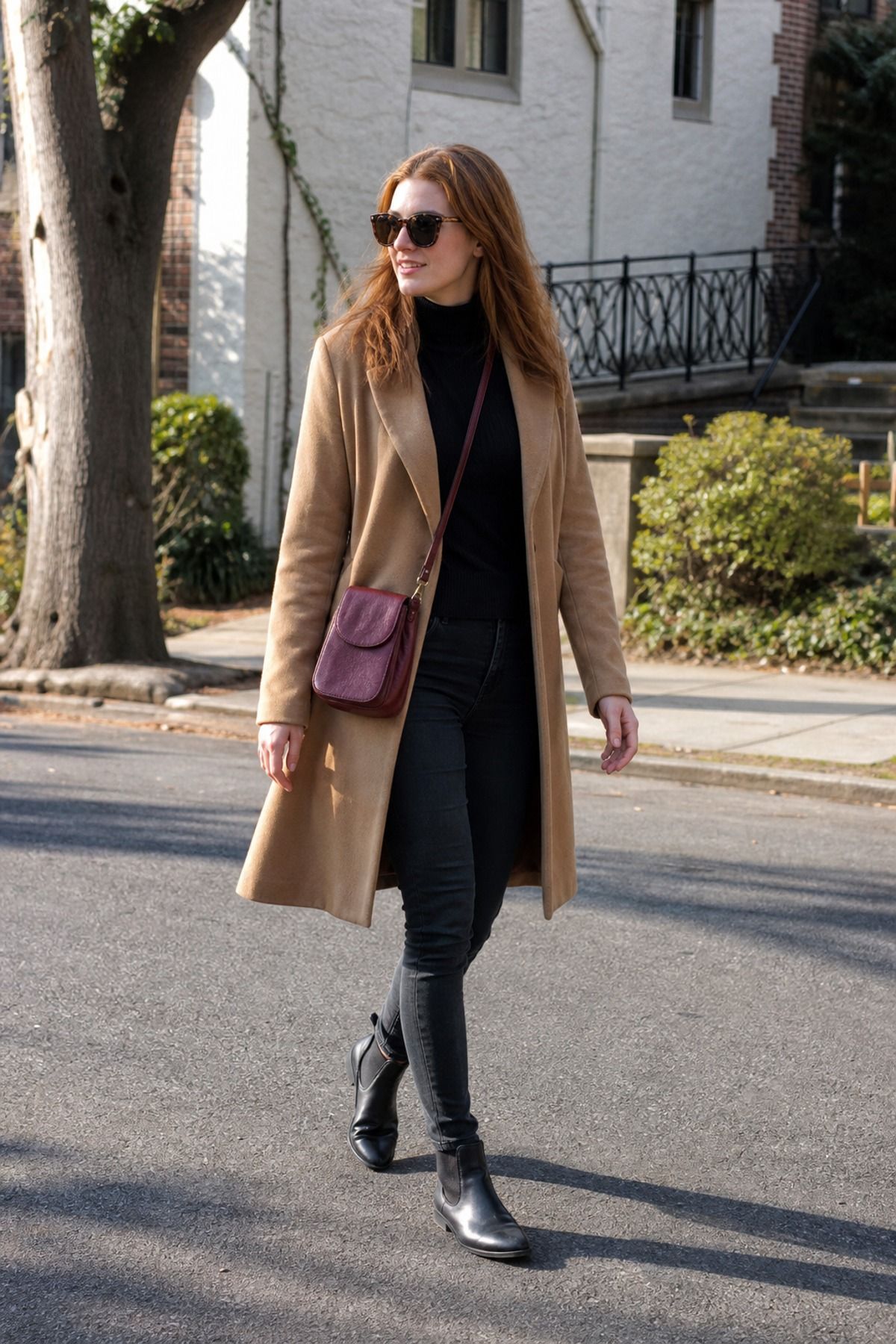 Woman in a long camel coat, black turtleneck, black skinny jeans and black ankle boots with a burgundy crossbody bag and sunglasses on a tree-lined street