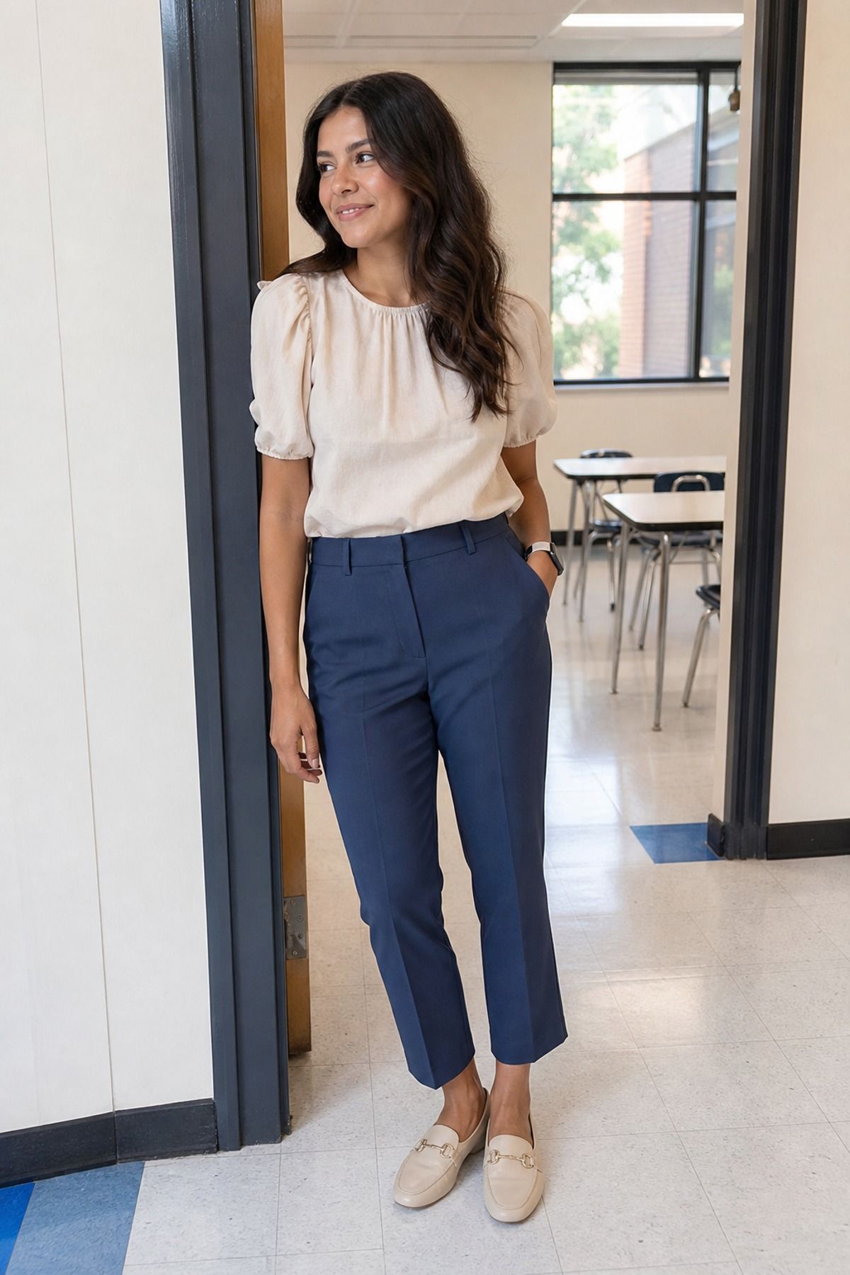 Woman in a cream puff-sleeve blouse, navy cropped trousers and beige loafers standing in a classroom doorway, a cute teacher outfit