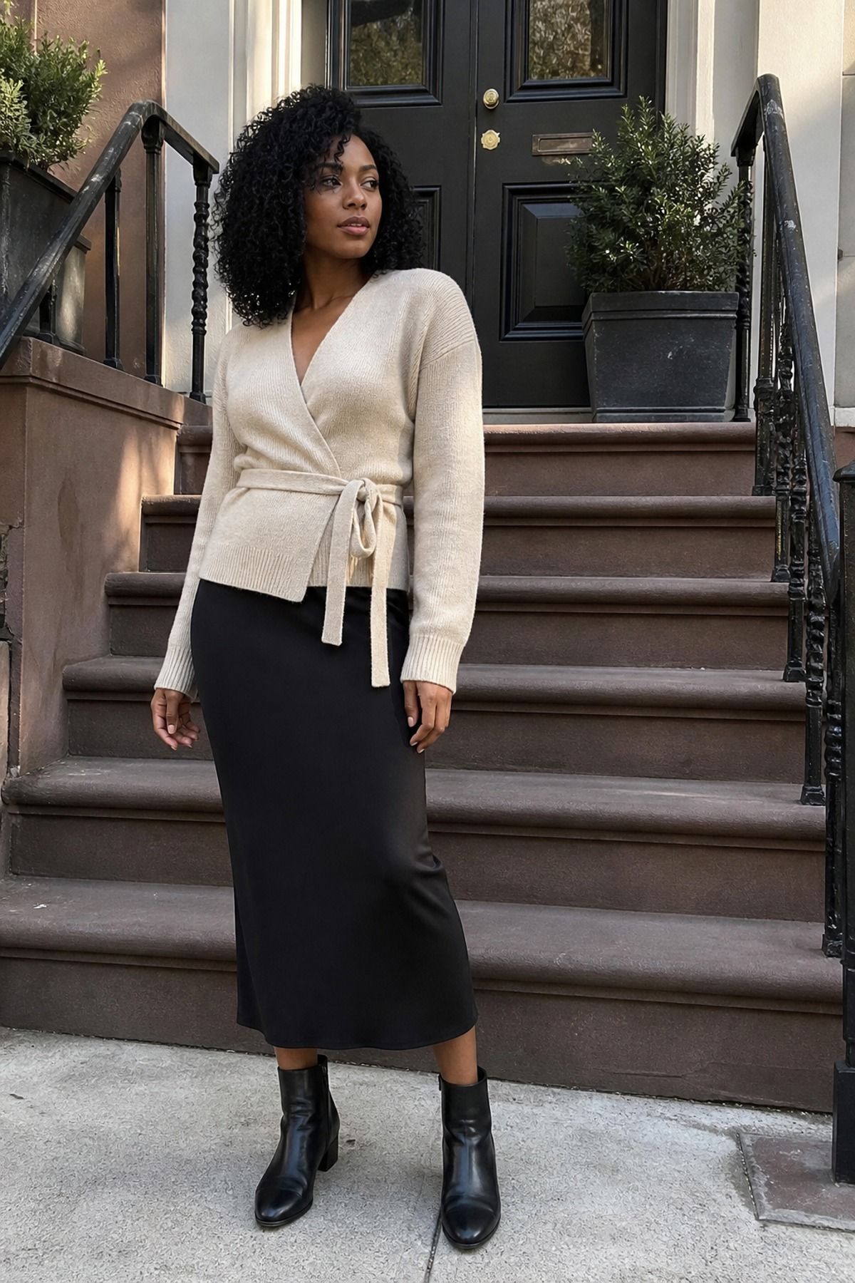 Woman in a cream wrap cardigan tied at the side, black column midi skirt and black lug ankle boots on brownstone steps