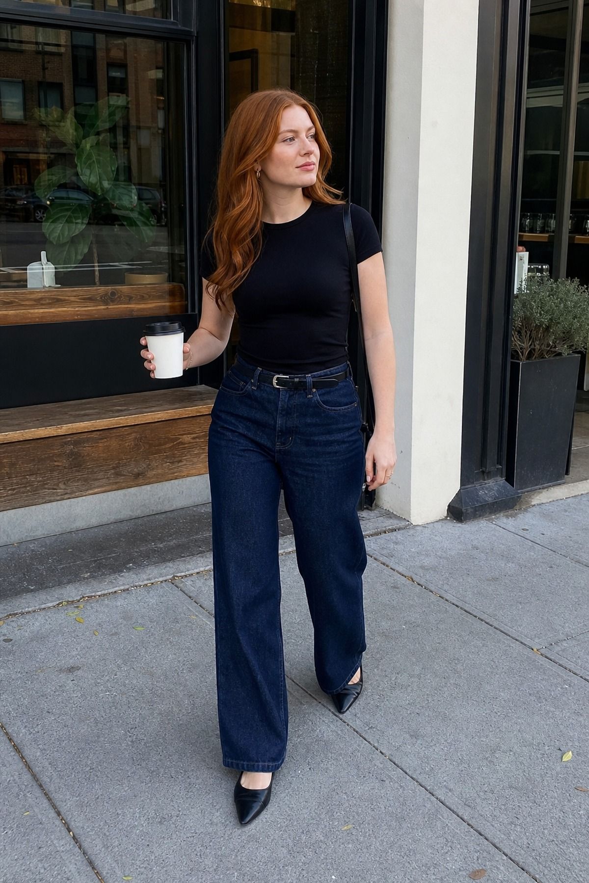 Woman in a black fitted tee, black belt, dark-wash wide trouser jeans and black pointed flats holding a coffee outside a cafe