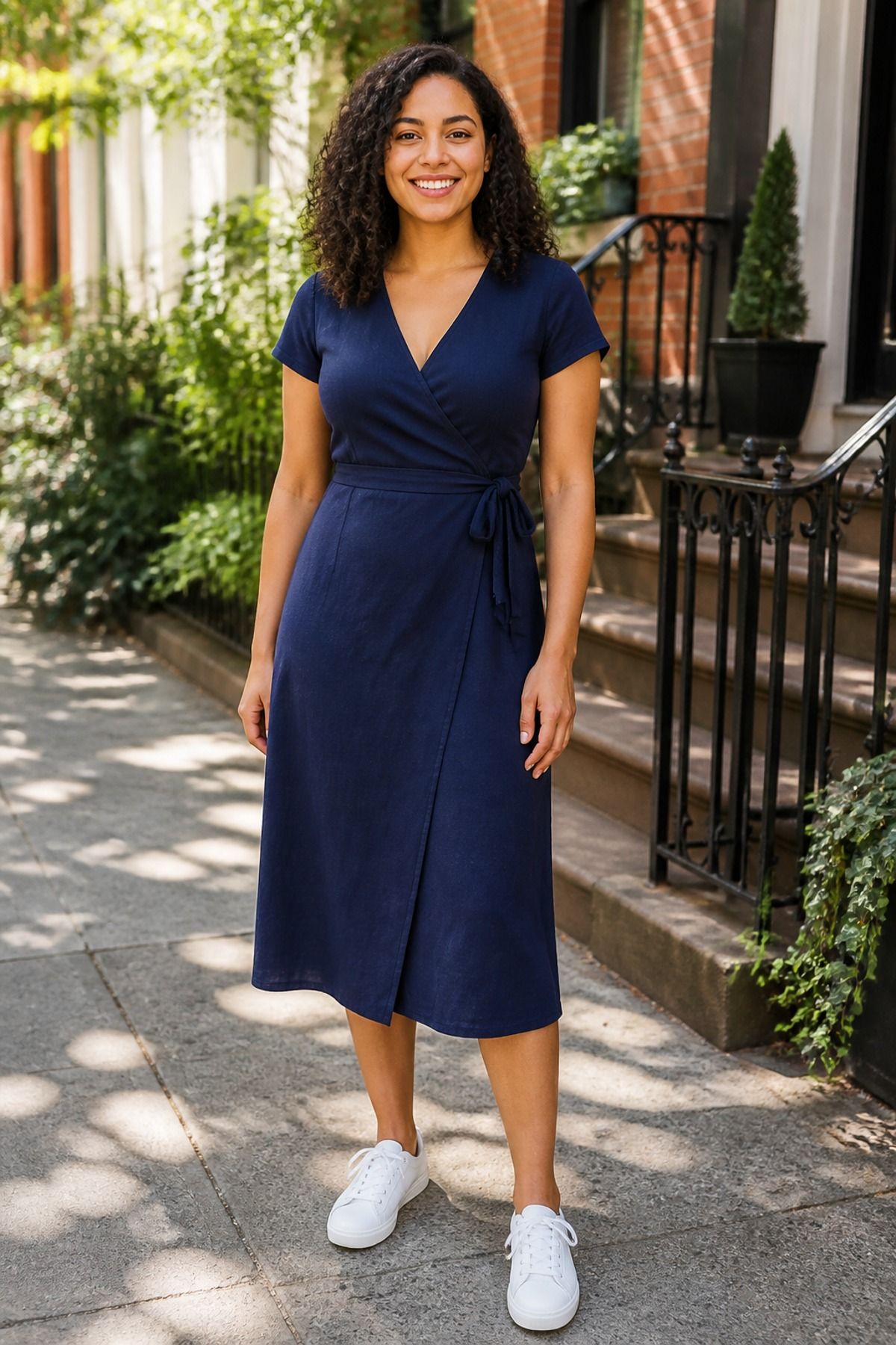 Woman in a navy wrap midi dress with short sleeves and white low sneakers on a sunny brownstone sidewalk
