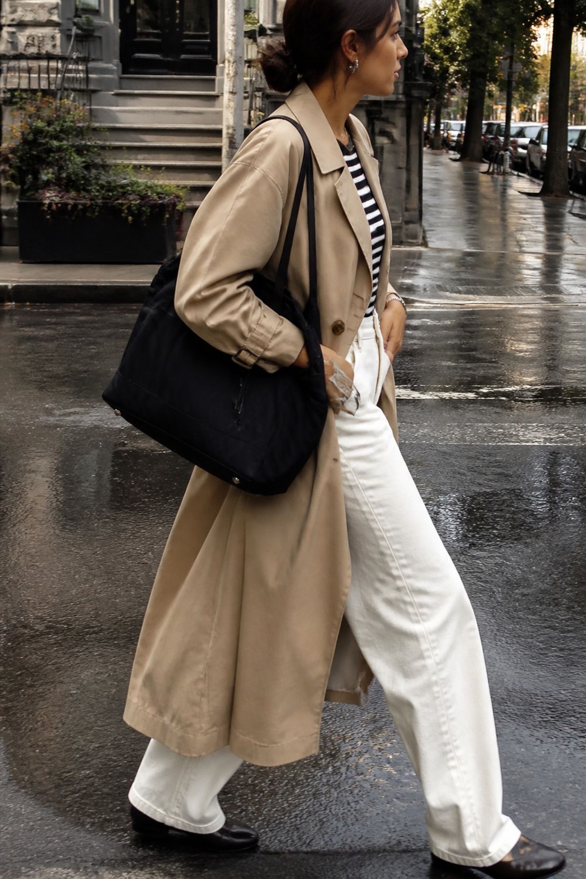 Woman in a beige trench coat, striped top, cream wide-leg trousers and black loafers carrying a black quilted nylon work bag on a wet street