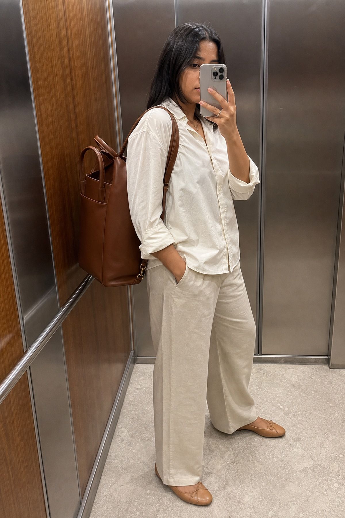 Elevator mirror selfie of a woman in a cream linen shirt and beige wide trousers wearing a brown leather convertible tote backpack