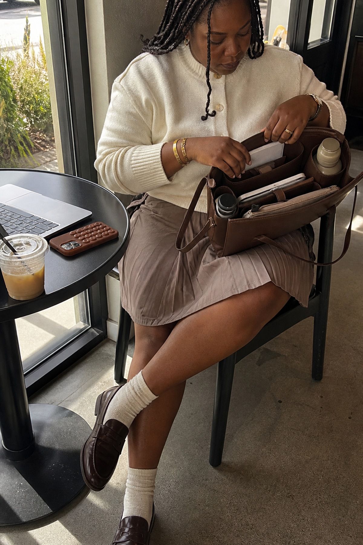 Woman in a cream sweater, taupe pleated skirt, white socks and brown loafers organizing an open work bag with compartments at a cafe table with a laptop