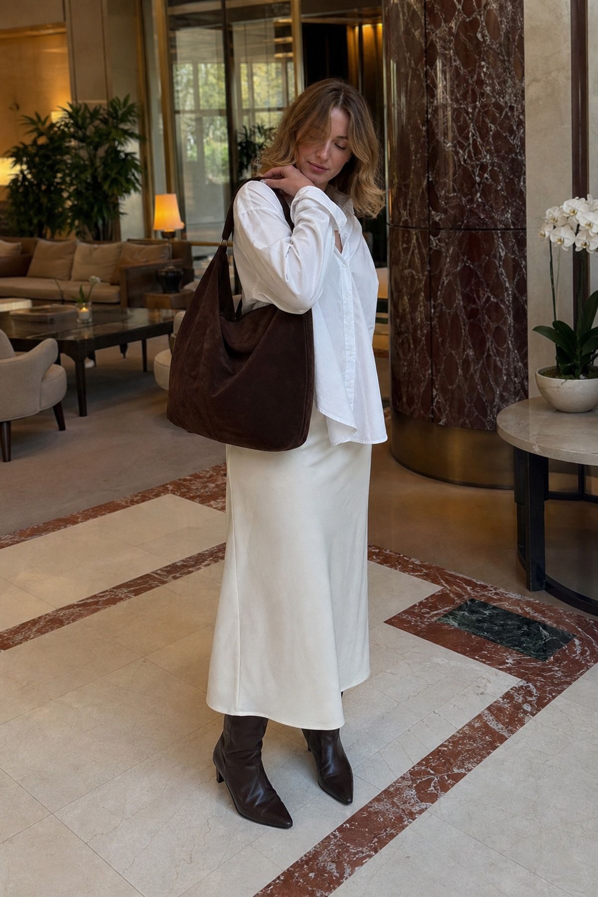 Woman in a white shirt, white satin midi skirt and dark brown pointed boots with a large dark brown leather shoulder bag in a hotel lobby
