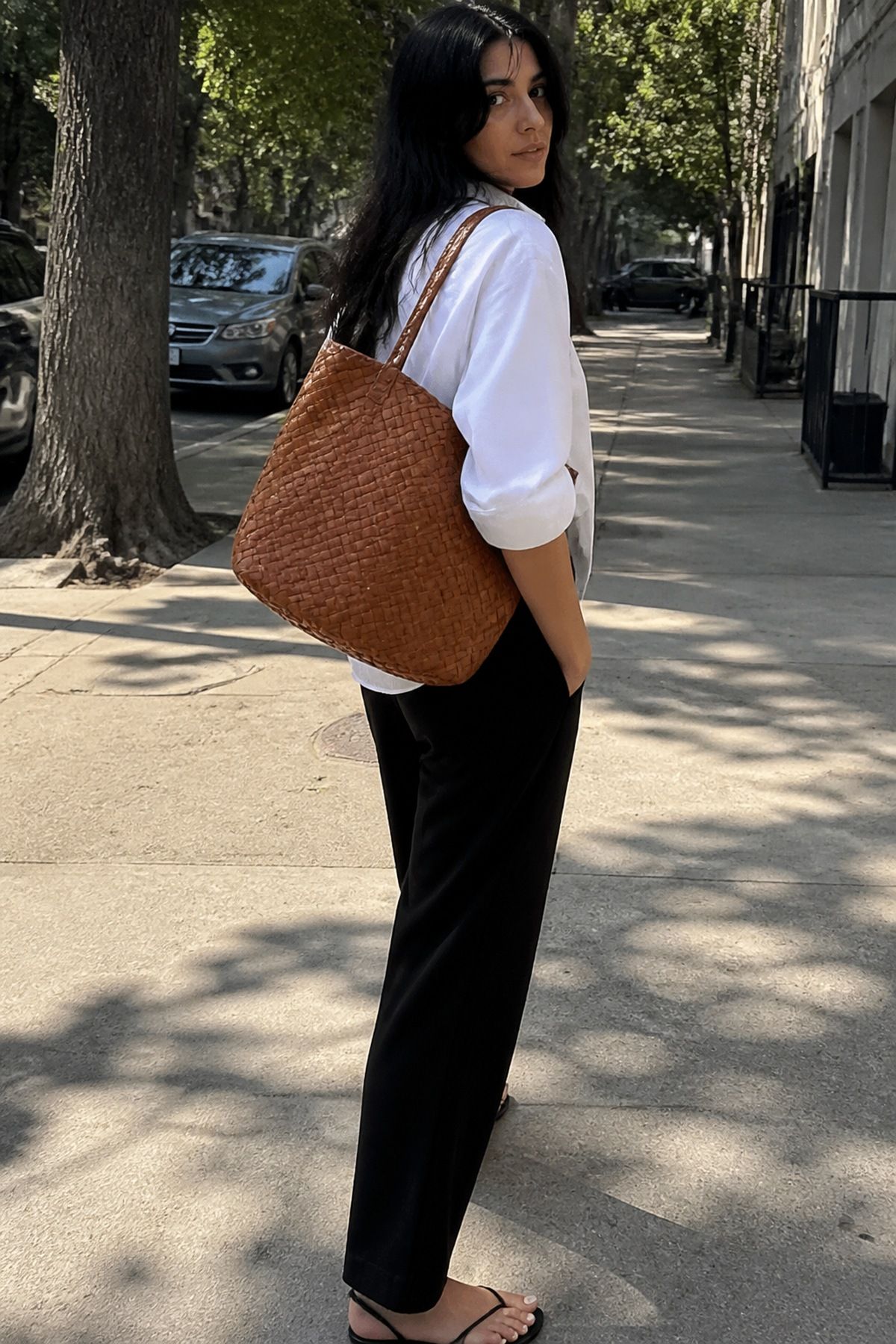 Back view of a woman in a white shirt and black trousers with a tan woven leather work tote on her shoulder on a sunny tree-lined street
