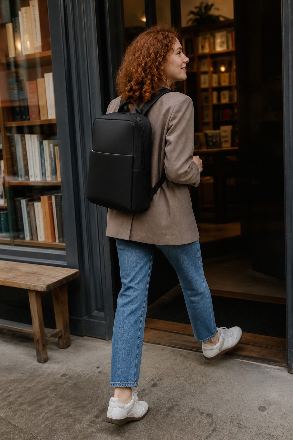 Back view of a woman in a taupe blazer, light-wash jeans and white sneakers wearing a sleek black work backpack while entering a bookshop