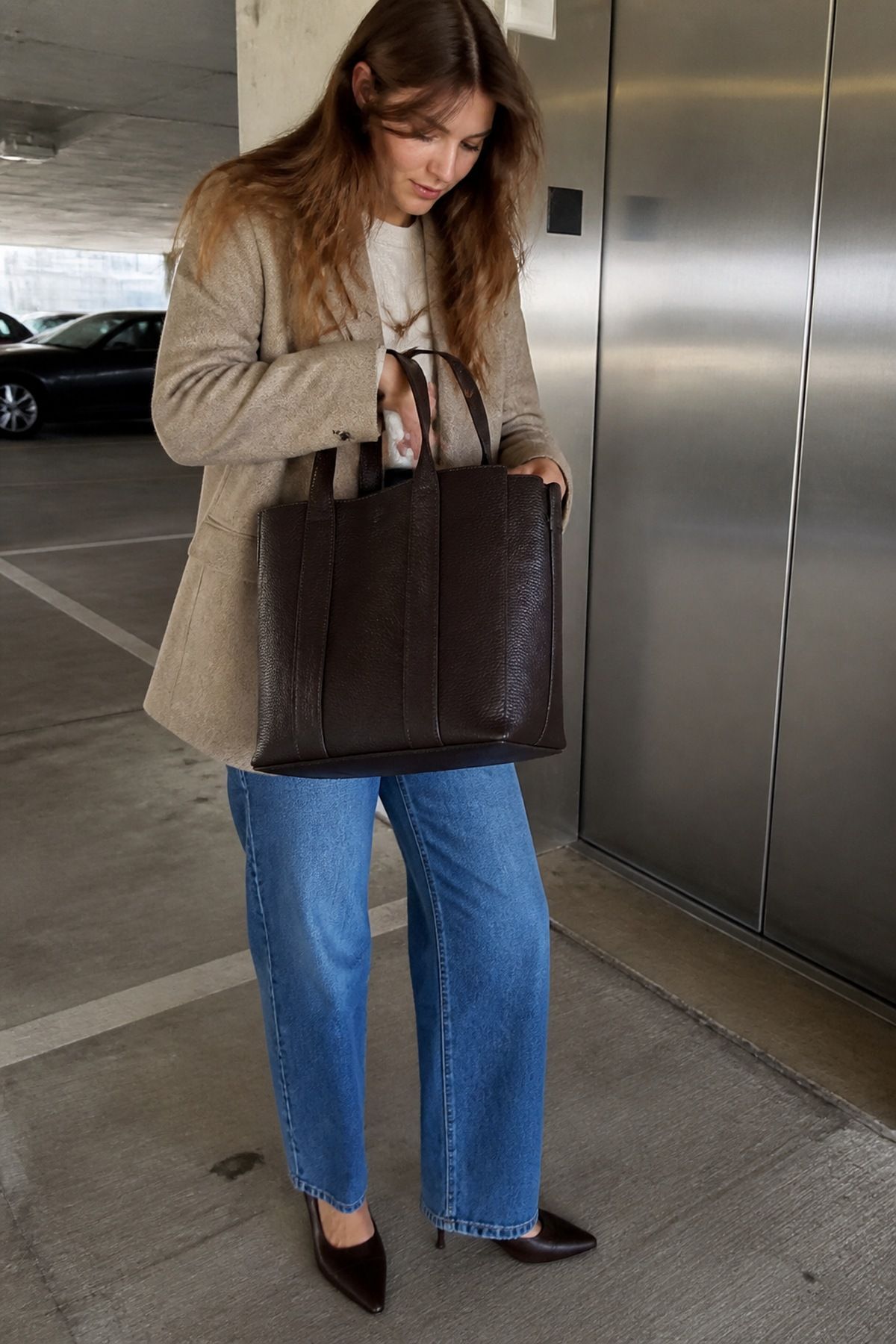 Woman in a beige blazer, gray tee, mid-blue wide-leg jeans and black pointed heels holding a large dark brown pebbled leather tote in a parking garage