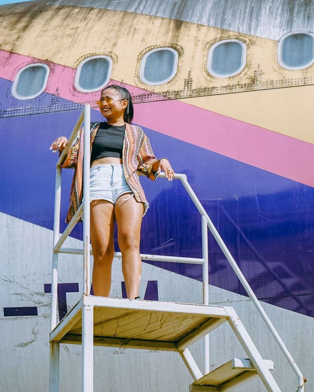 Evan Kristine smiling on white airplane stairs in a striped kimono, crop top and denim shorts
